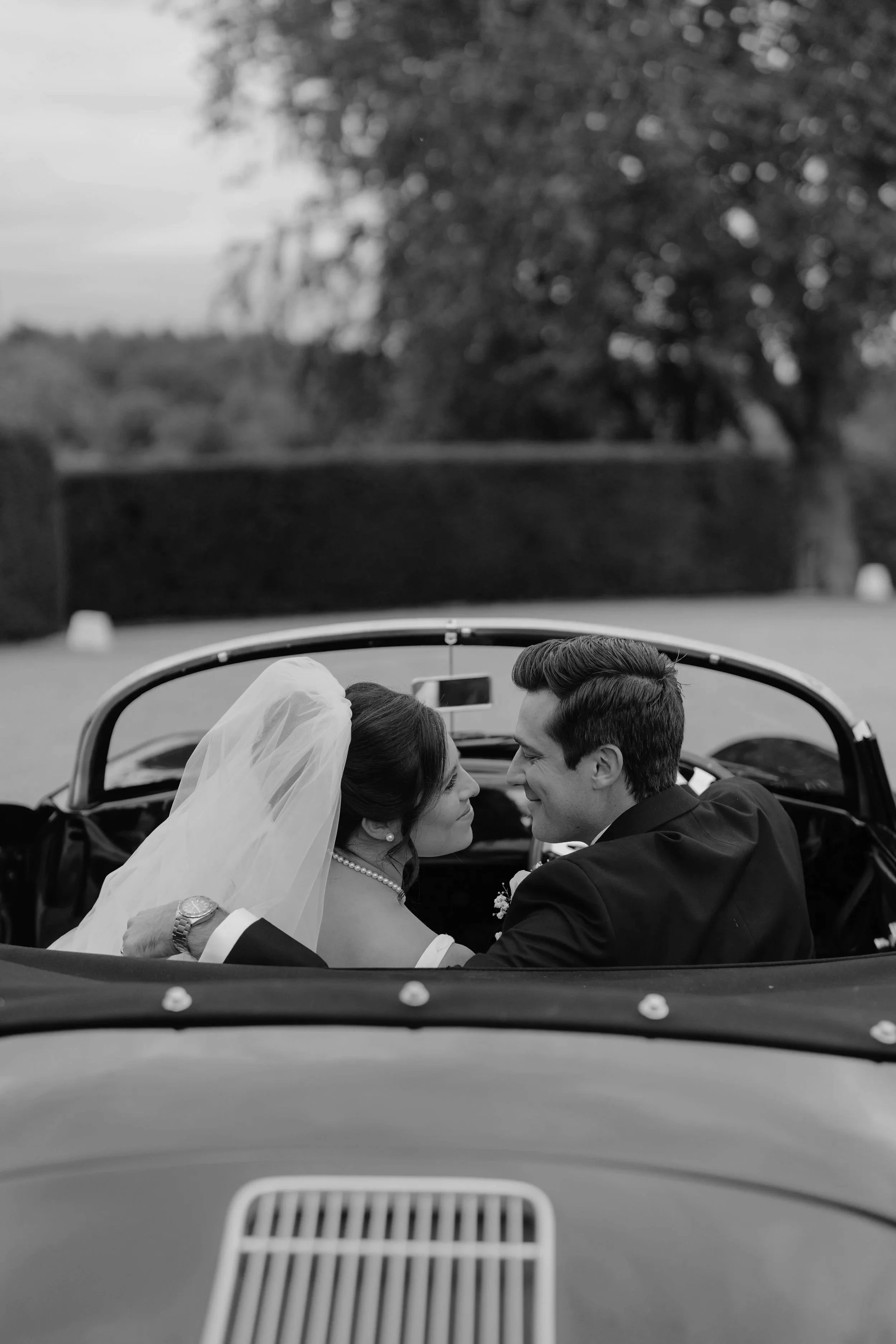 A black and white photograph of a bride and groom sitting in a vintage convertible car, facing each other closely, with a large tree and open landscape in the background.