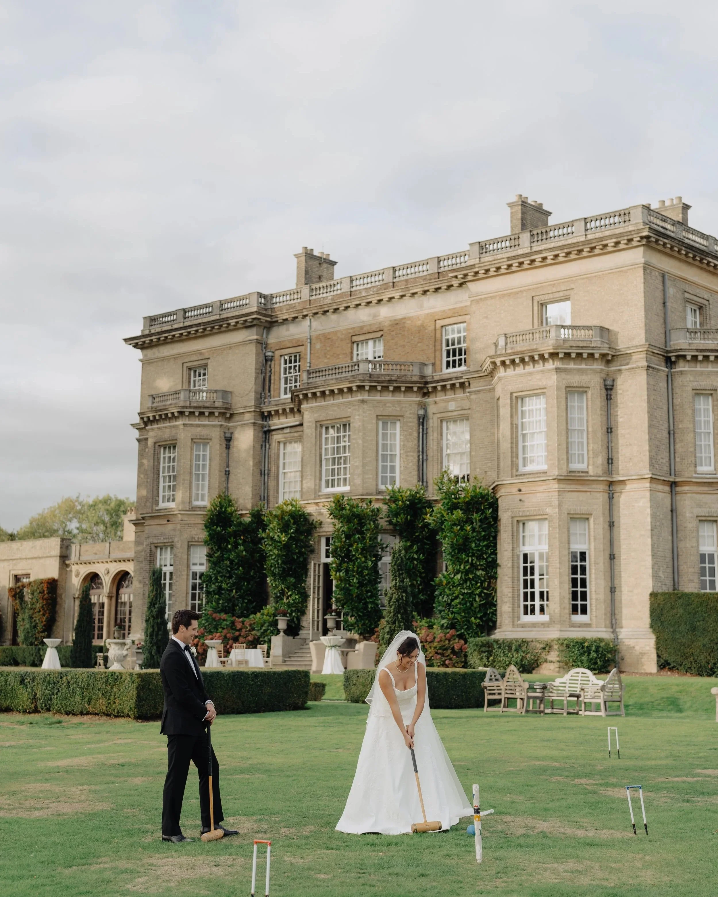 A bride and groom playing croquet on the lawn of a grand historic building with large windows and decorative stonework.