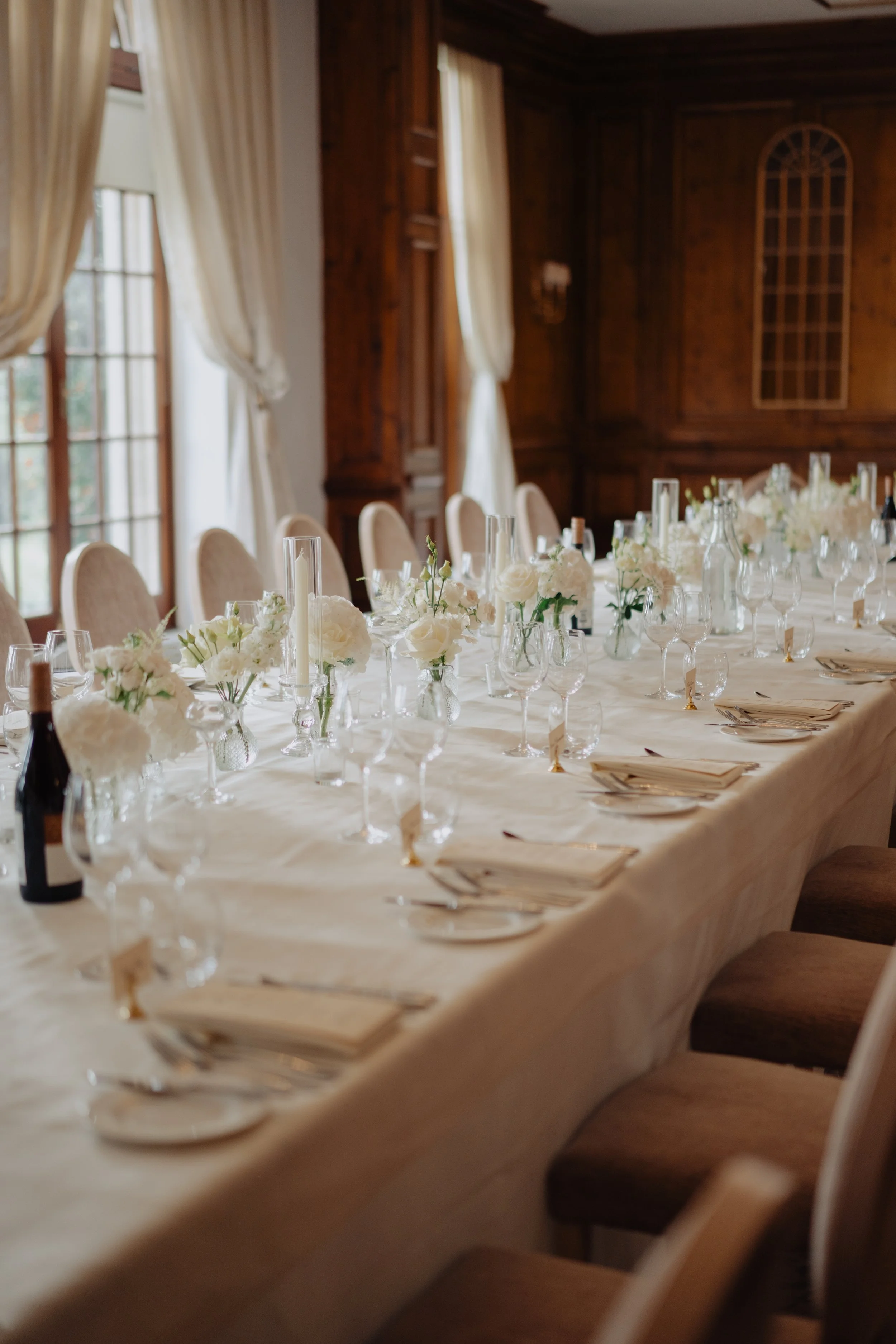 Elegant dining table set with white floral centerpieces, candles, and glassware in a wood-paneled room with large windows and cream curtains.