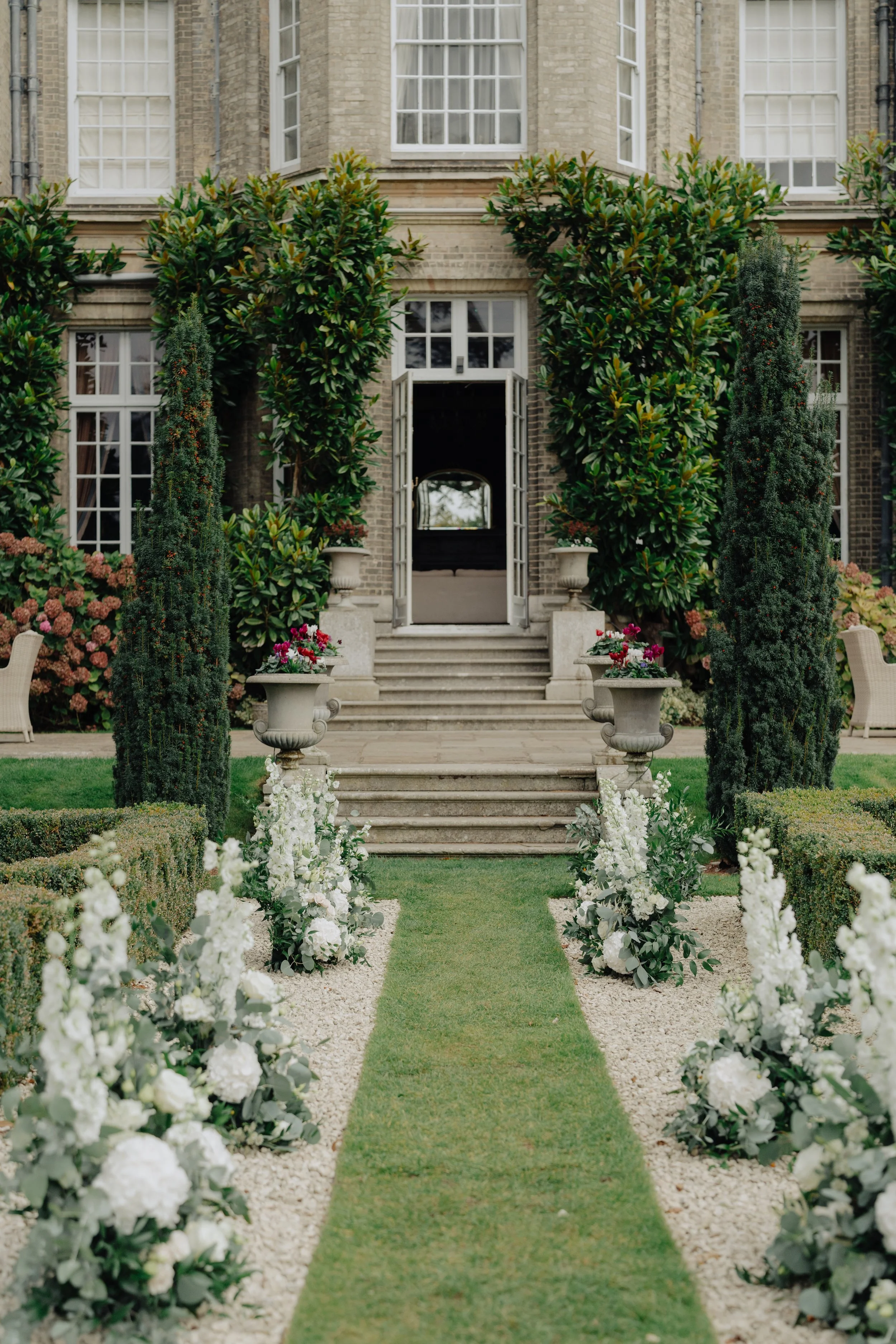 A garden with a central pathway covered in grass, flanked by rows of white flowers and small hedge bushes, leading to a building with large windows and open front doors, surrounded by tall green trees.