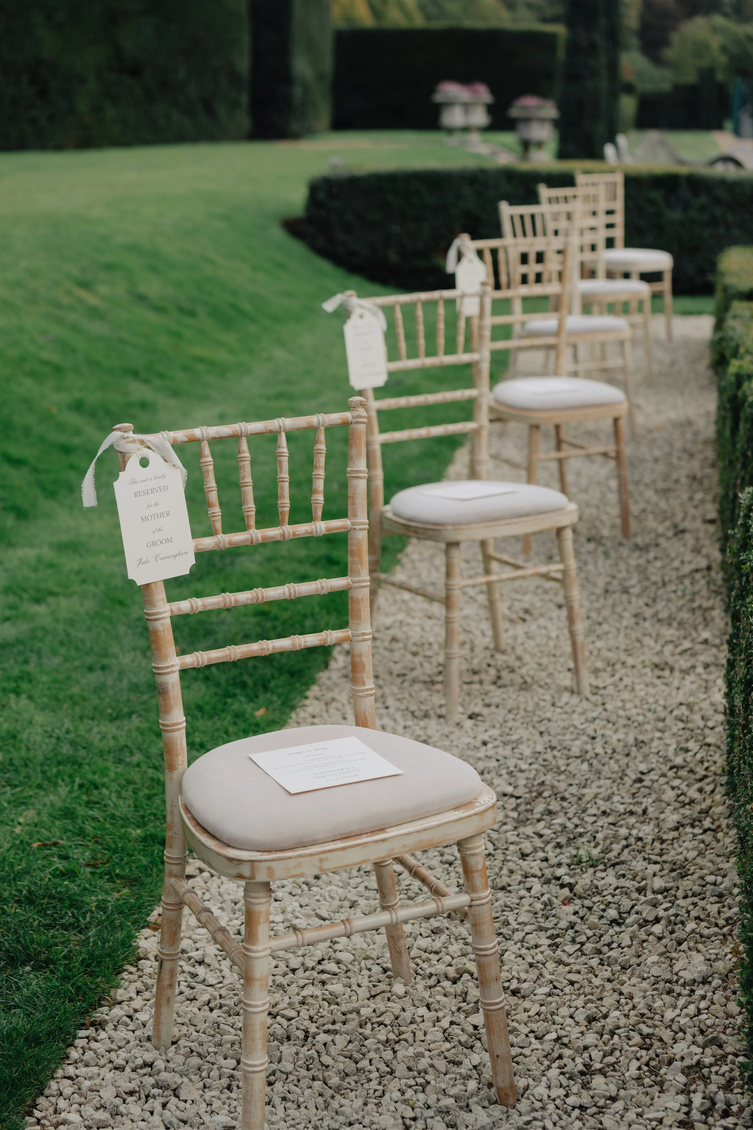 A row of elegant wooden chairs with white cushions and reserved signs for a wedding ceremony on a gravel pathway in a garden setting.