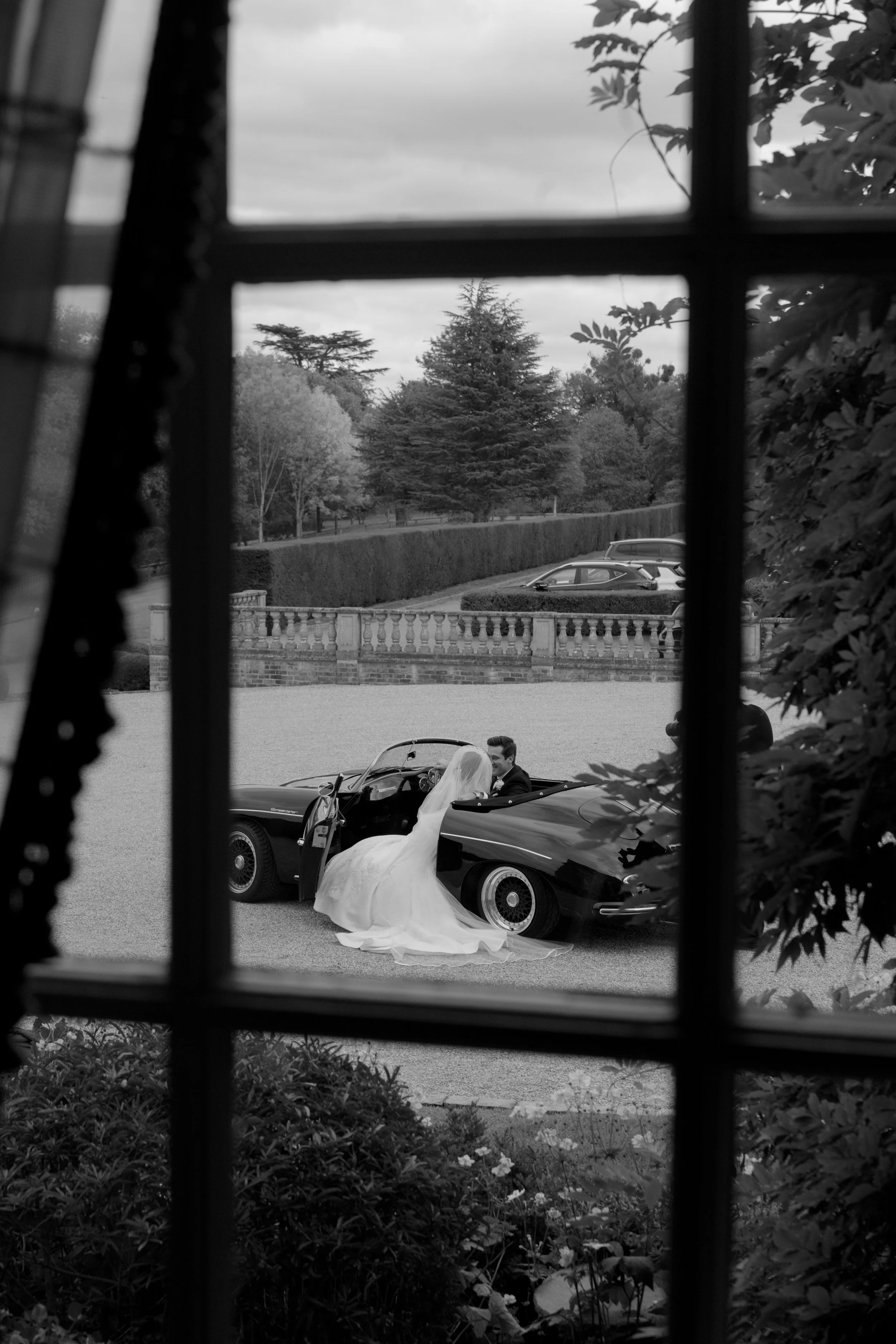A black and white photograph of a bride and groom in a vintage convertible car, seen through a window with a frame, outdoors on a cloudy day with trees and parked cars in the background.
