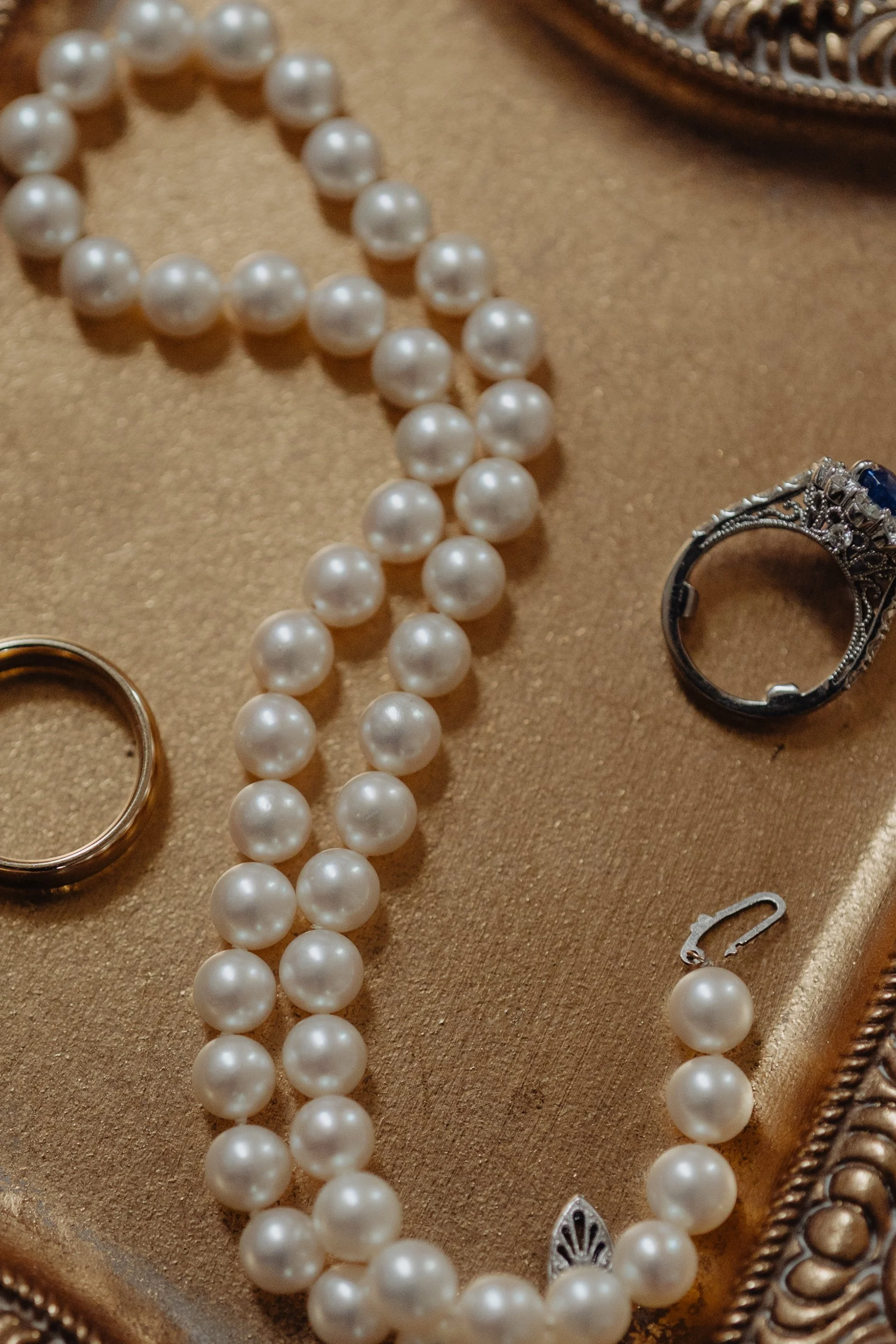A string of white pearl necklaces, a silver ring, a blue gemstone ring, and a small safety pin placed on a gold-colored jewelry tray.