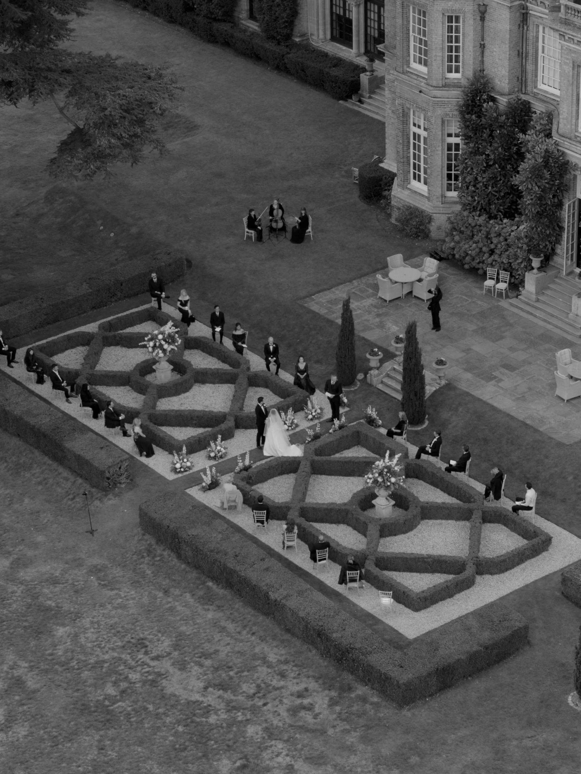 Black and white aerial view of a wedding ceremony in a garden with a hedge maze, wedding couple, guests, and chairs, near a large mansion.