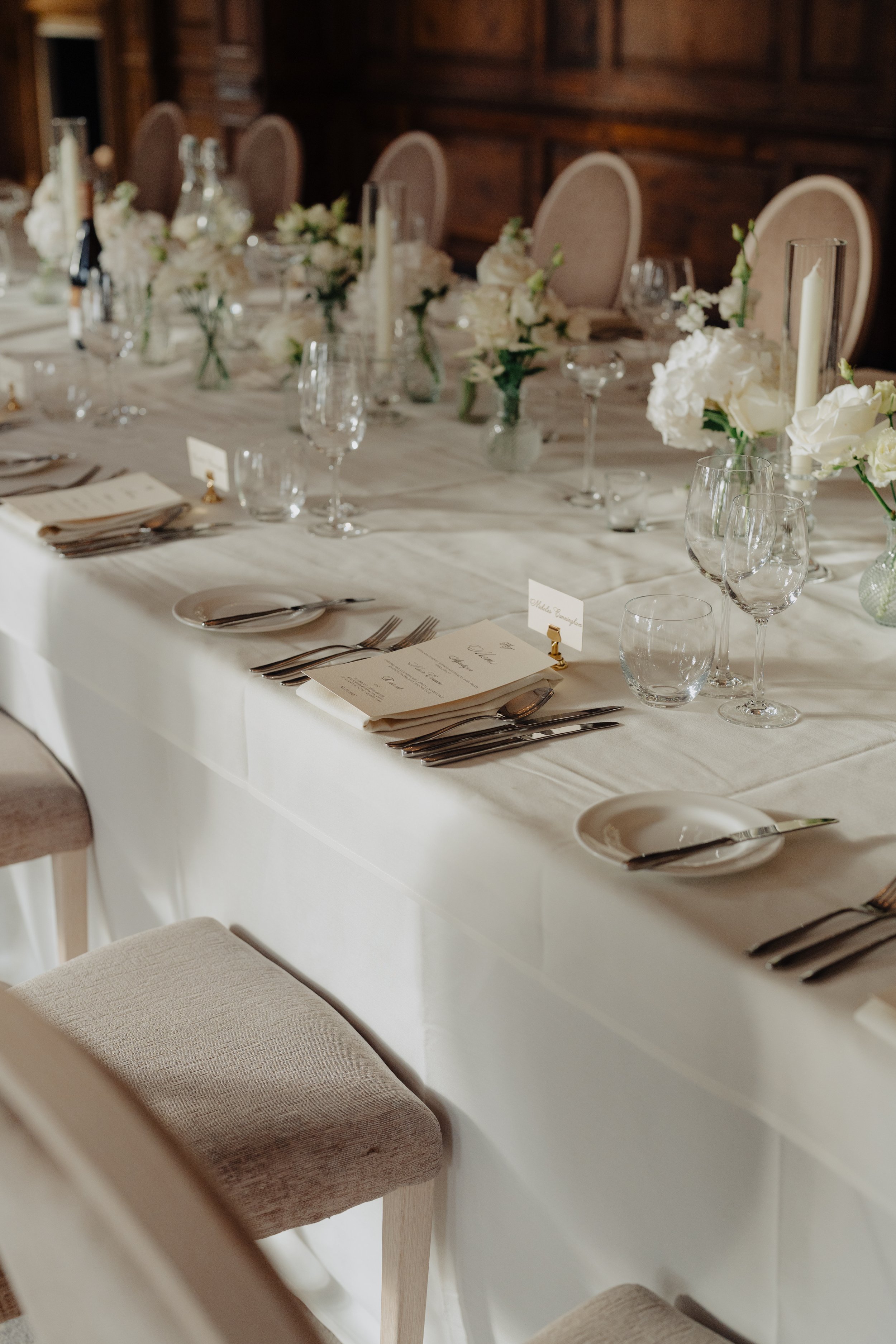 A long dining table set for a formal event with white tablecloth, multiple glassware, silverware, and floral centerpieces, surrounded by cream-colored chairs with high backs.