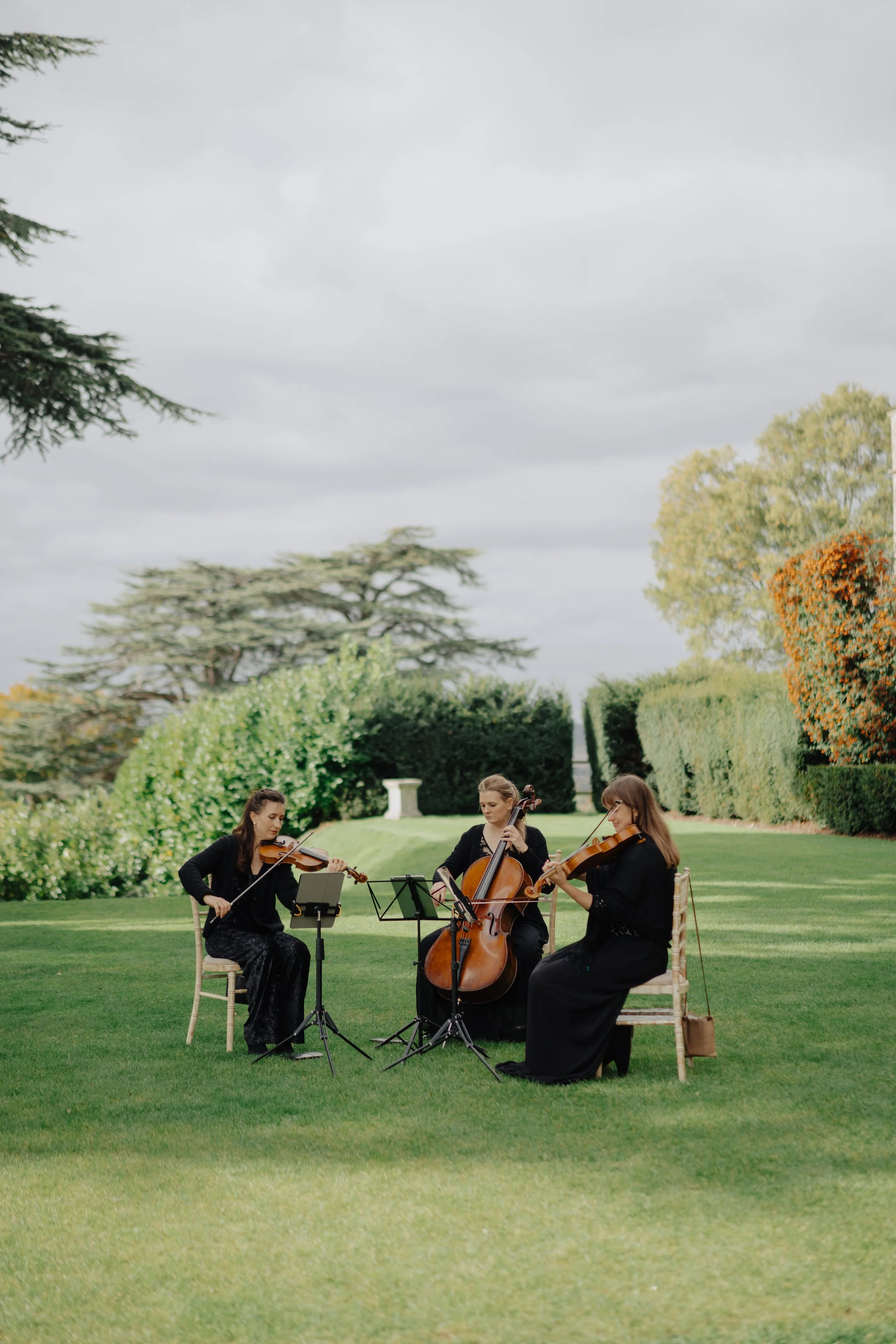 Three women dressed in black performing classical music outdoors, sitting on chairs on a well-maintained lawn, with music stands in front of them, surrounded by bushes, trees, and a cloudy sky.