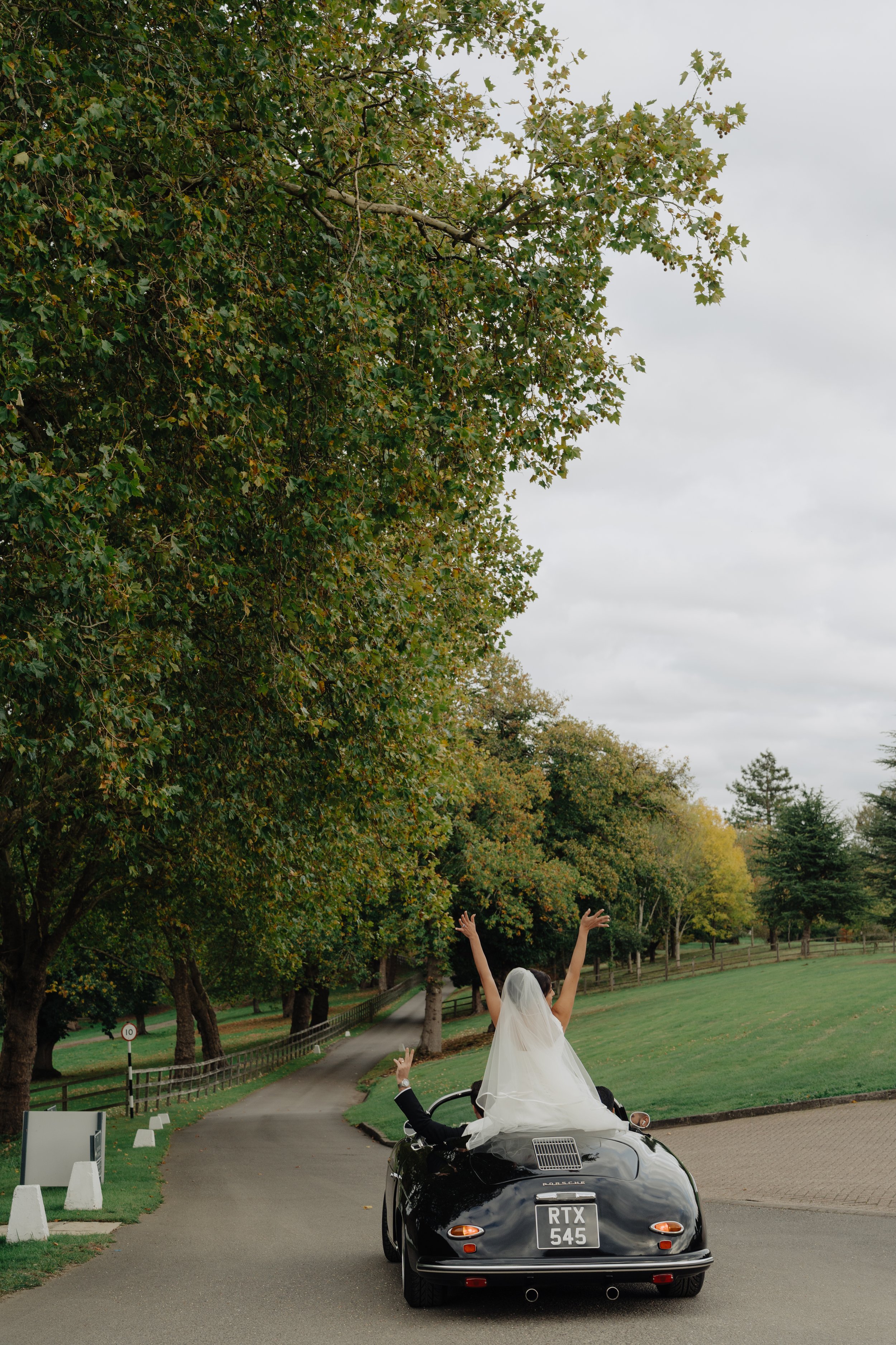 A black Porsche sports car with a bride sitting on its hood, wearing a white wedding dress and veil, raising her arms. A groom in a tuxedo is sitting behind her, reaching out to her. The background includes a park with lush green grass, trees, and a 