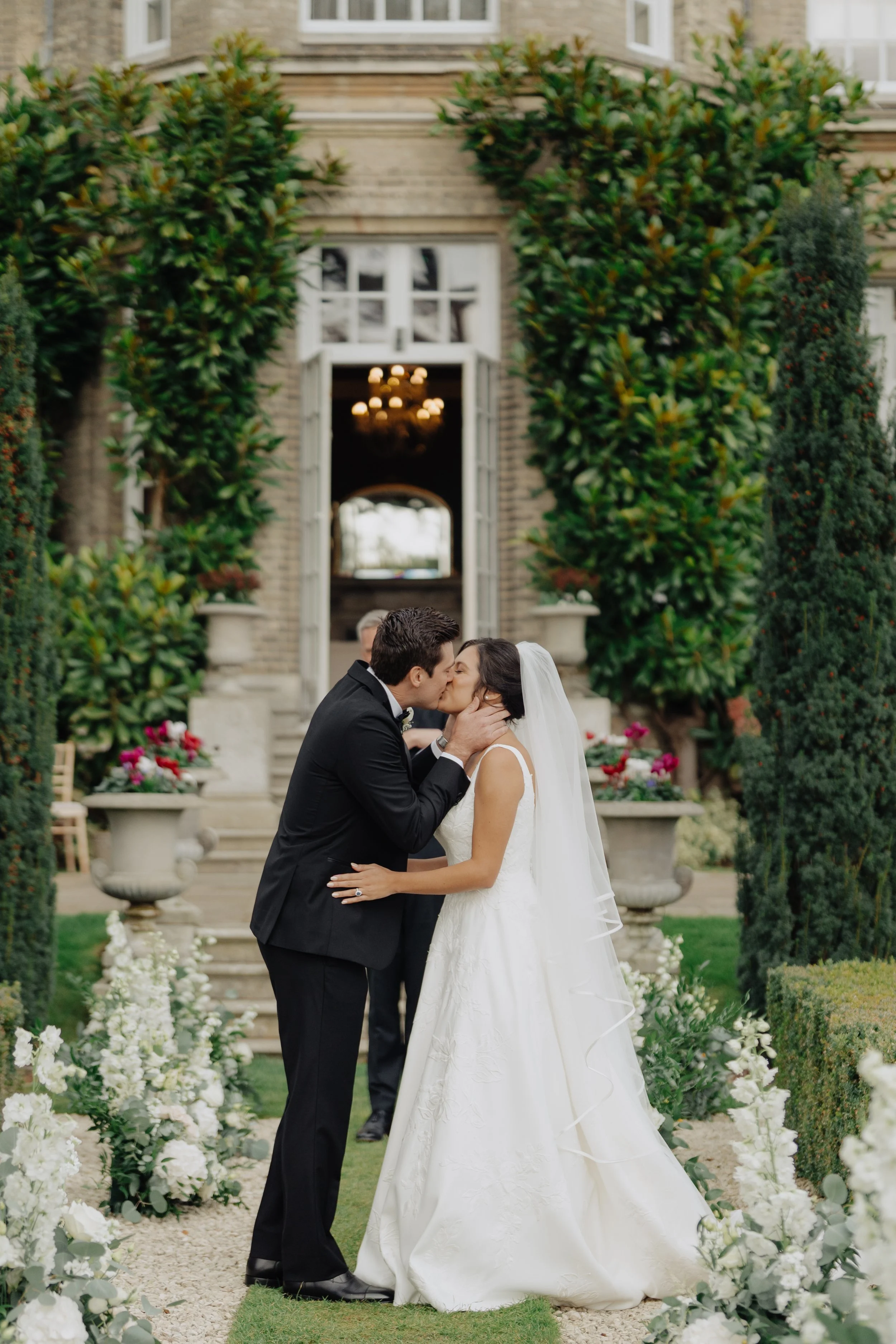 A bride and groom kiss during their wedding ceremony outdoors in front of a historic building with large green bushes and white flowers surrounding them.