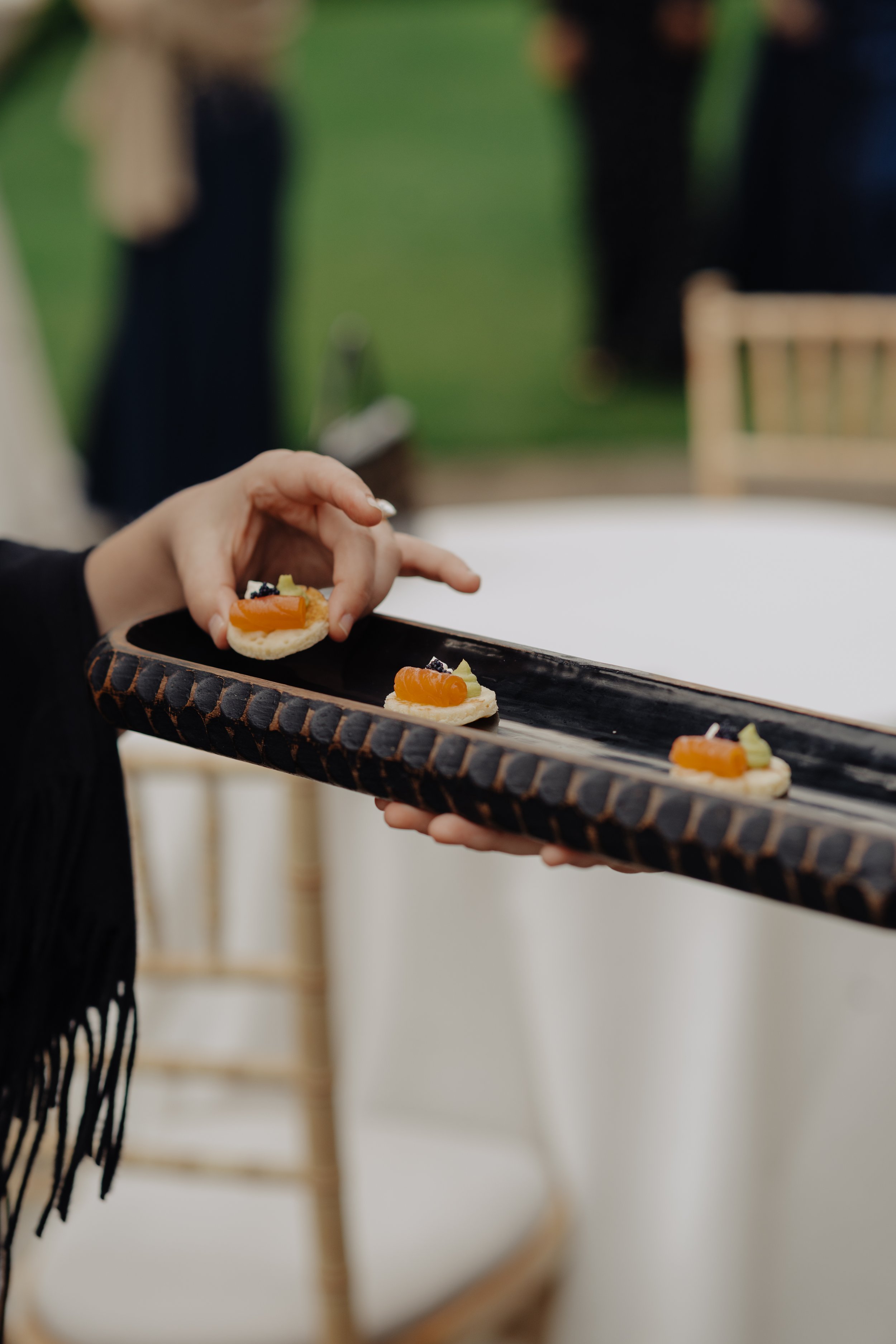 Person holding a black tray with small appetizers topped with radish, vegetable, and seafood items at an indoor event.