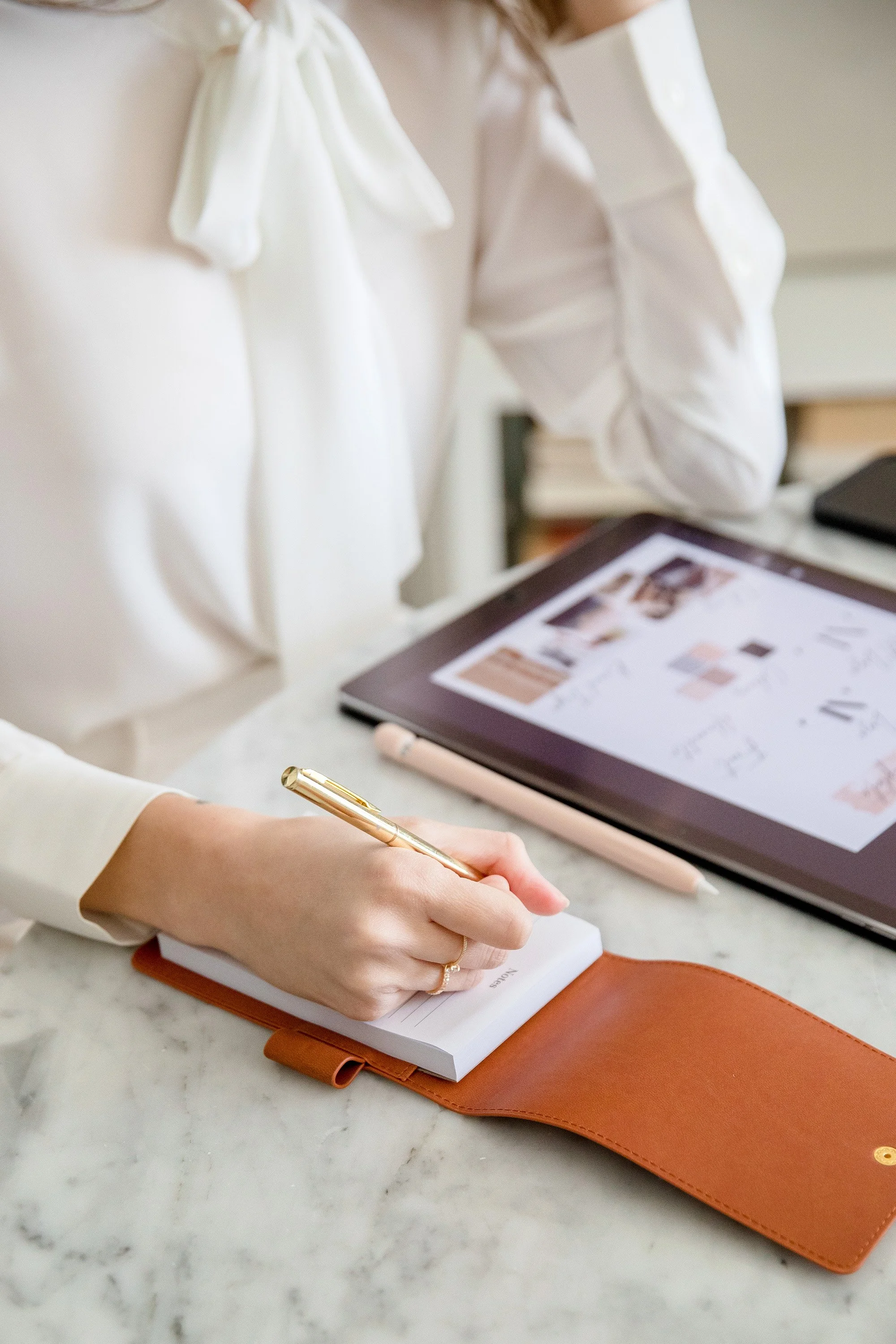 A person writing in a small notepad with a gold pen, resting on a brown leather cover on a white marble table, with a tablet displaying a website in the background.