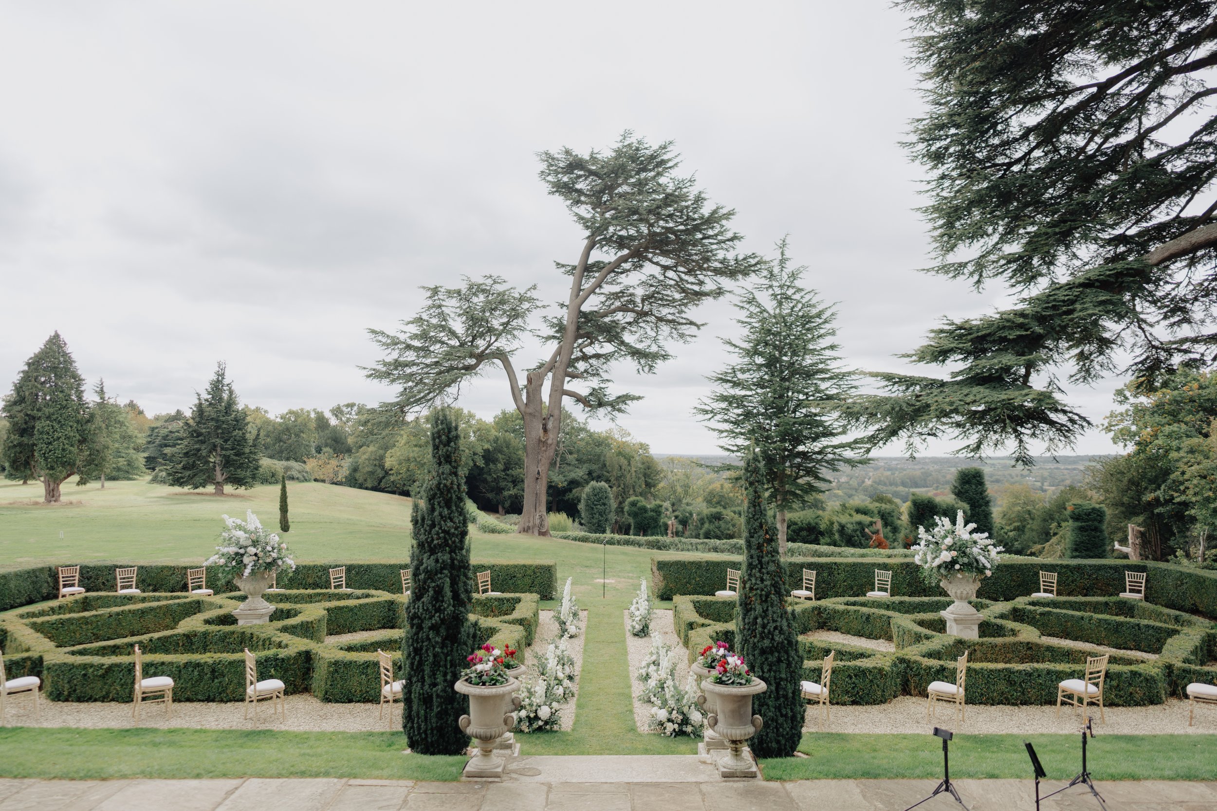 A landscaped outdoor wedding venue with chairs arranged in two sections, floral arrangements in large vases, trimmed hedges, and tall trees under an overcast sky.