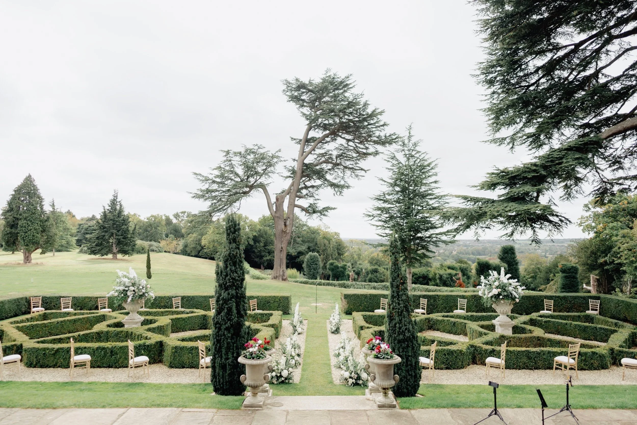 Outdoor wedding venue with neatly trimmed hedges, flower arrangements, and white chairs set up for guests. Tall trees and a cloudy sky in the background.