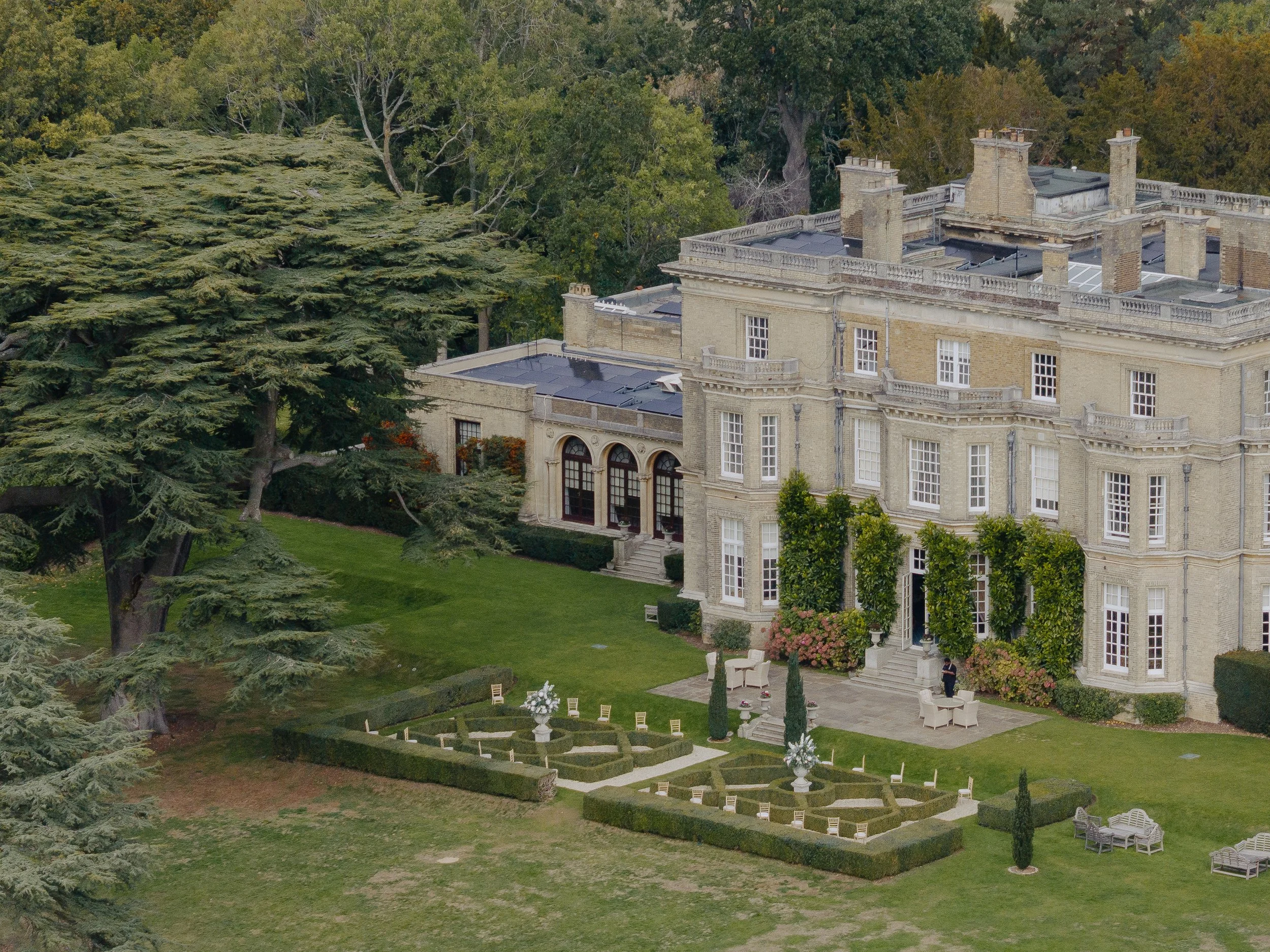 A large historic mansion with beige stone walls and multiple chimneys, surrounded by lush green trees and a well-manicured garden with flower beds, white chairs, and outdoor tables.