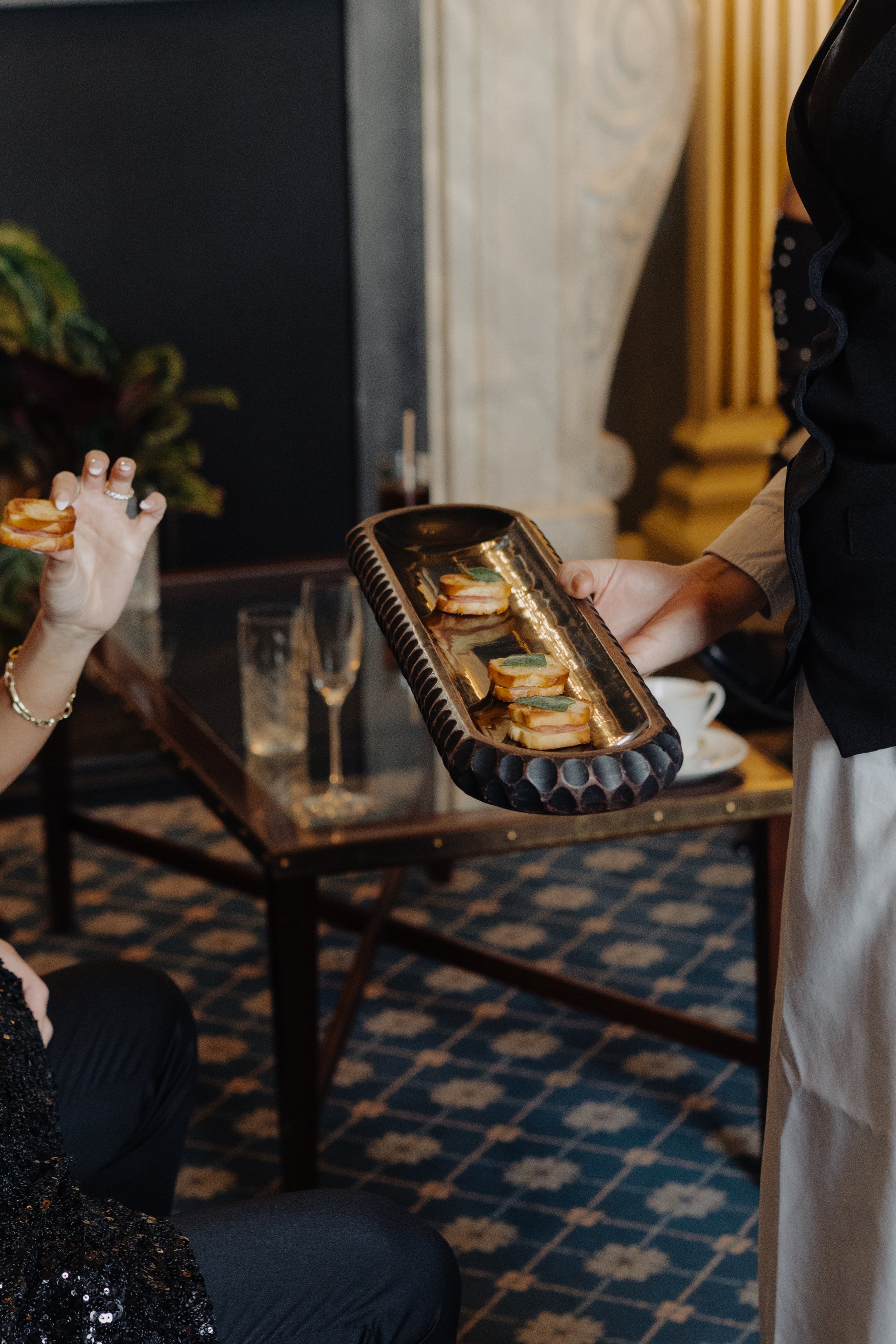 A person in formal attire is serving a plate with small sandwiches to a guest, while another guest is reaching to take a sandwich from her tray. The setting appears to be a formal event or gathering, with a table, glassware, and decorative elements i