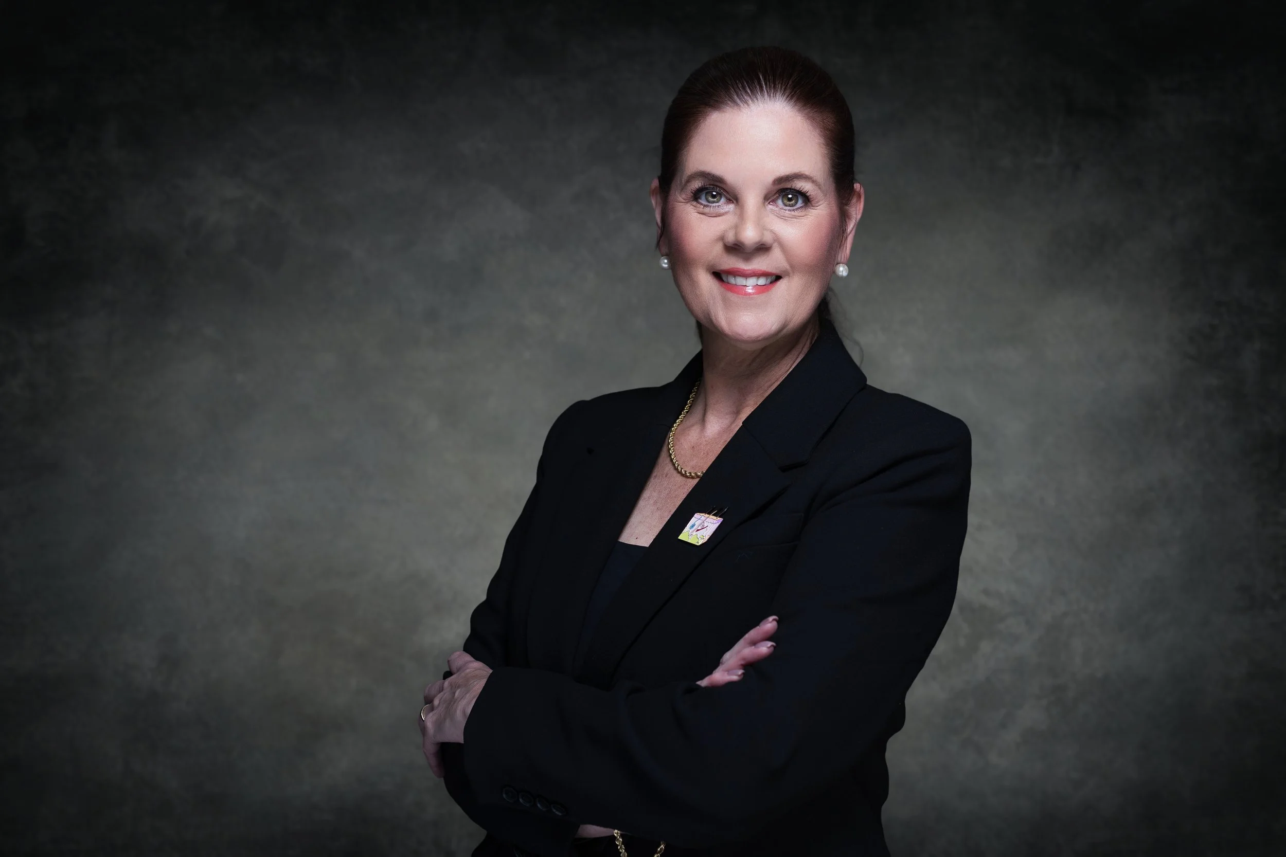 A professional woman with red hair, wearing a black suit and pearl earrings, smiling and standing with arms crossed against a dark background.
