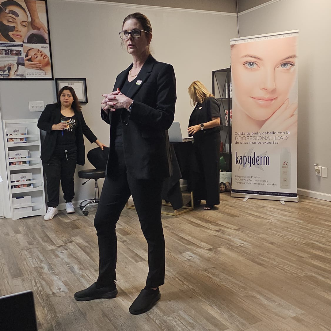 A woman in black business attire with glasses speaking or presenting, standing in a room with a wooden floor. Two other women are in the background, one holding a drink and the other working on a laptop, near a promotional banner for skincare products.