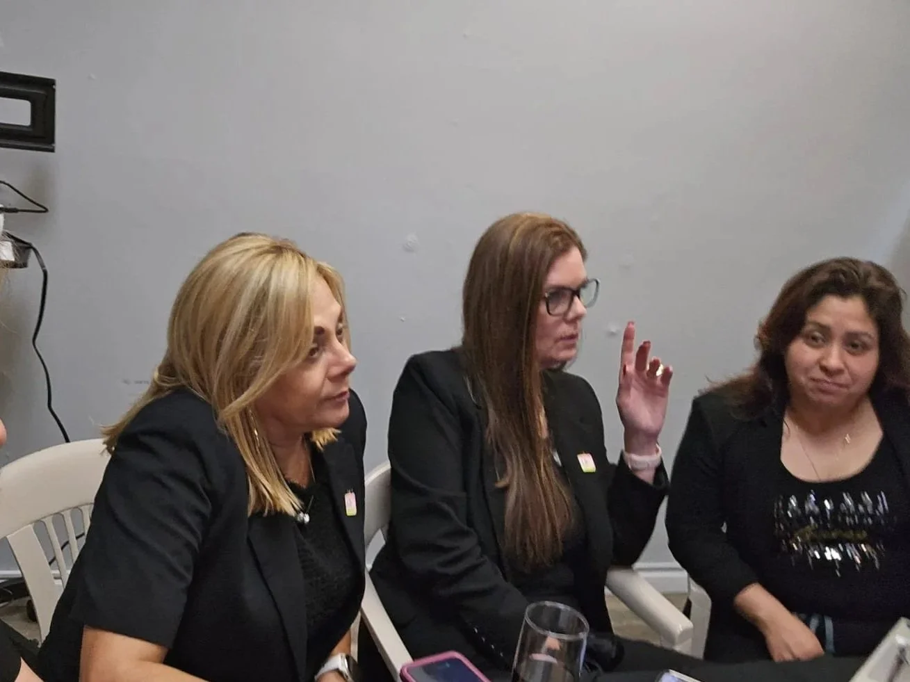 Three women seated at a table, engaged in conversation, with a plain wall in the background.