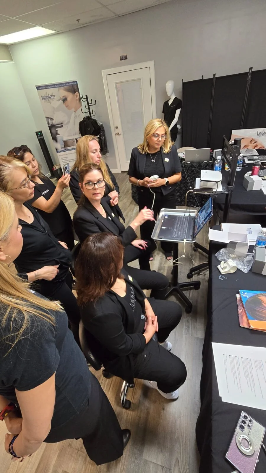 Several women gathered around a woman seated at a table, giving a presentation or demonstration with a laptop and computer monitors in a commercial or conference room.