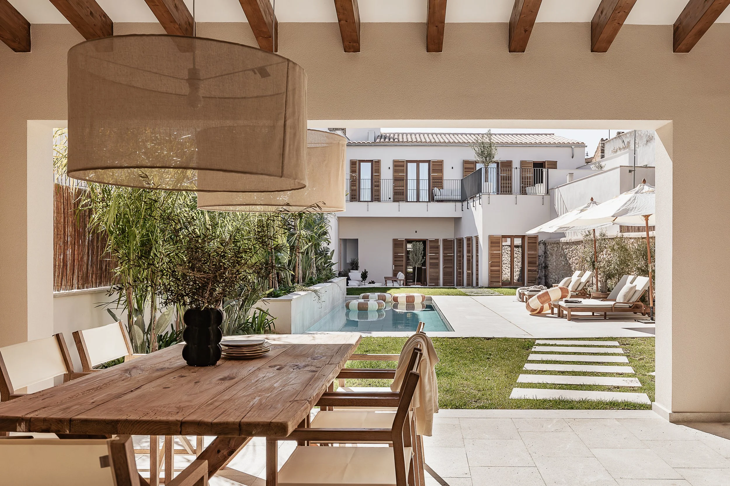Blick von einem Innenbereich auf einen Innenhof mit Pool und Terrassenbereich, umgeben von weißen Gebäuden mit Holzfenstern und -läden. Im Vordergrund ein Holz Esstisch mit einem Lampenschirm und Geschirr, im Hintergrund Liegestühle und Sonnenschirme am Pool.