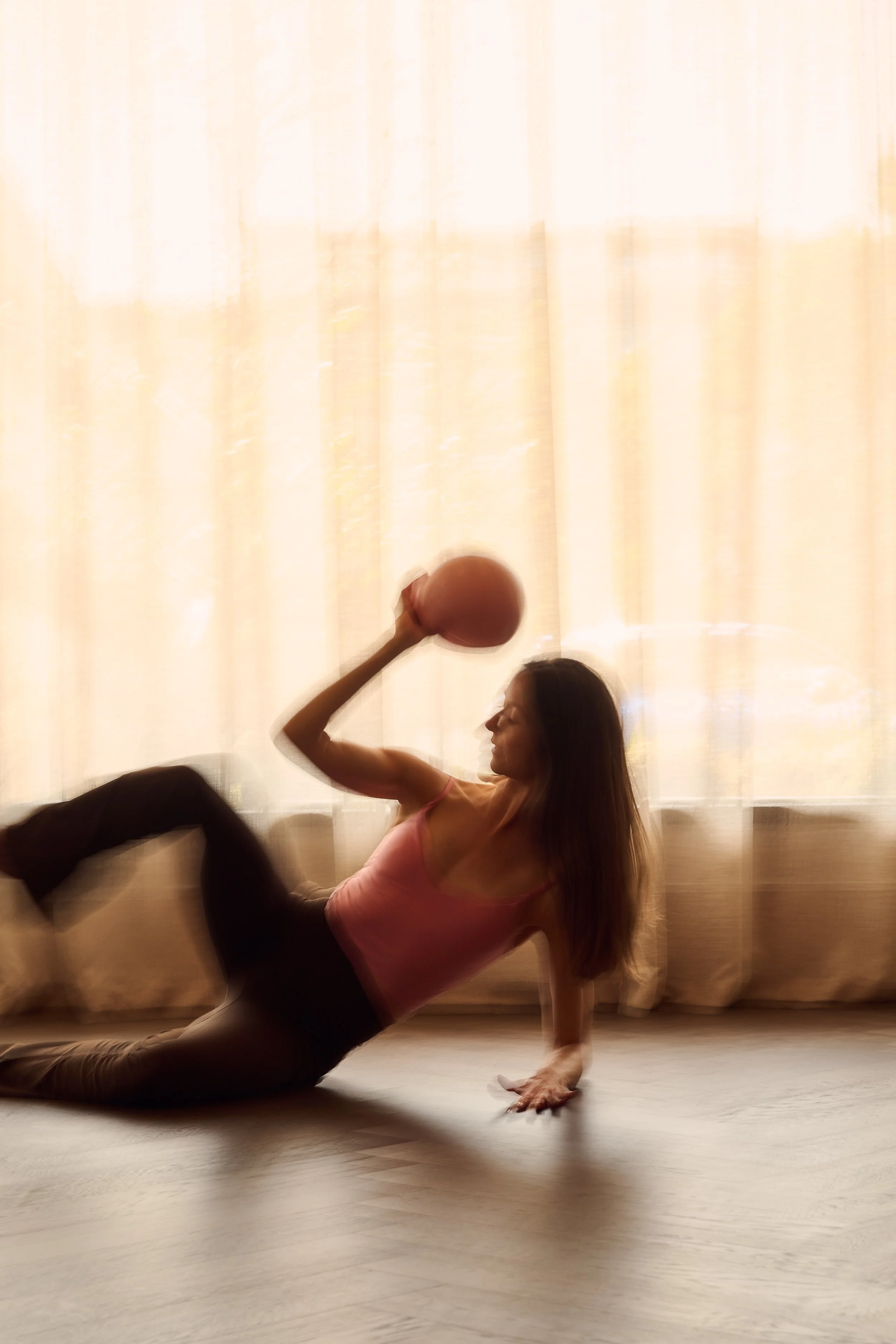 A woman performing a yoga pose on a light-coloured wooden floor in front of a beige curtain. She is holding a squishy ball with her right hand while supporting herself on her left hand and left leg, with her right leg bent and raised in the air.