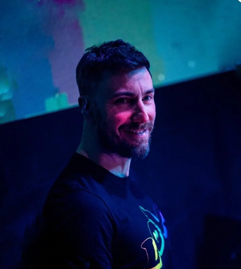 Uomo sorridente con barba e capelli corti in un ambiente con luci colorate, casual con maglietta nera.