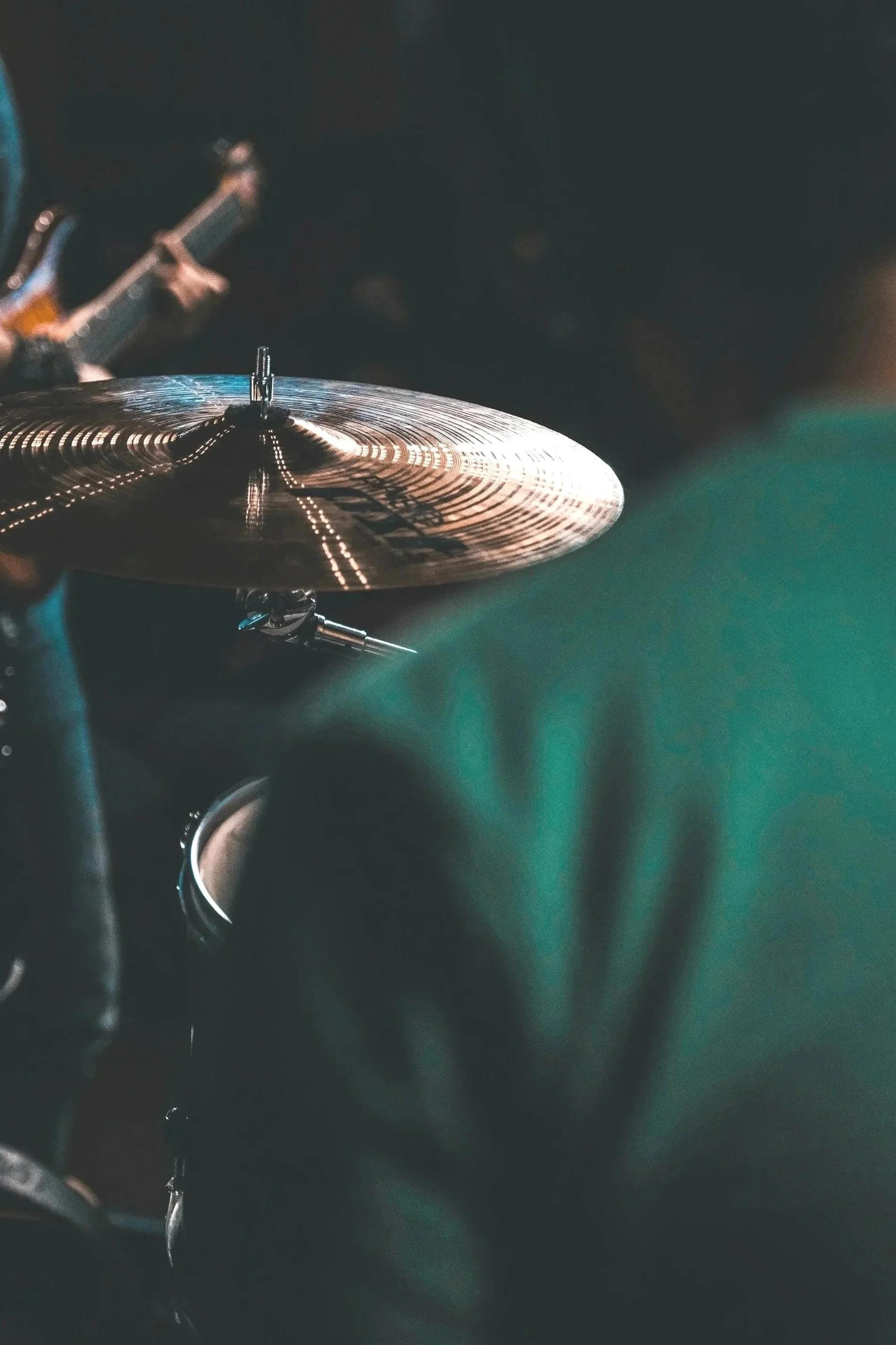 Particolare di un piatto di batteria, con un piatto in primo piano, e un musicista con chitarra sfocato sullo sfondo.