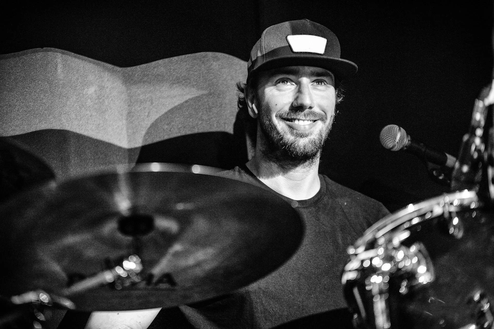 Un uomo sorridente con cappellino seduto dietro una batteria in uno studio di registrazione.