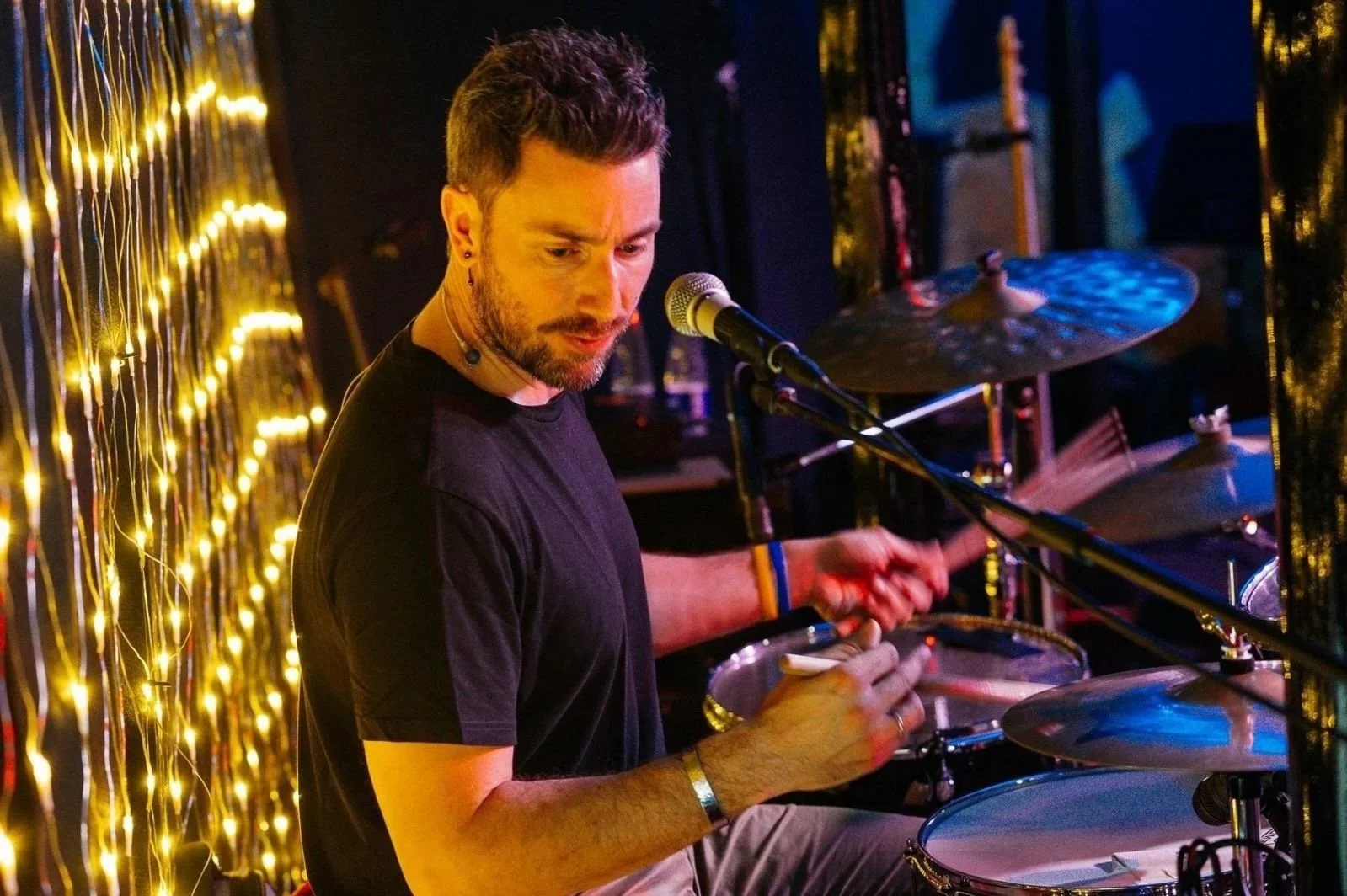 Un musicista con capelli corti e barba, che suona la batteria in un locale con luci calde e sfondo decorato con luci scintillanti.
