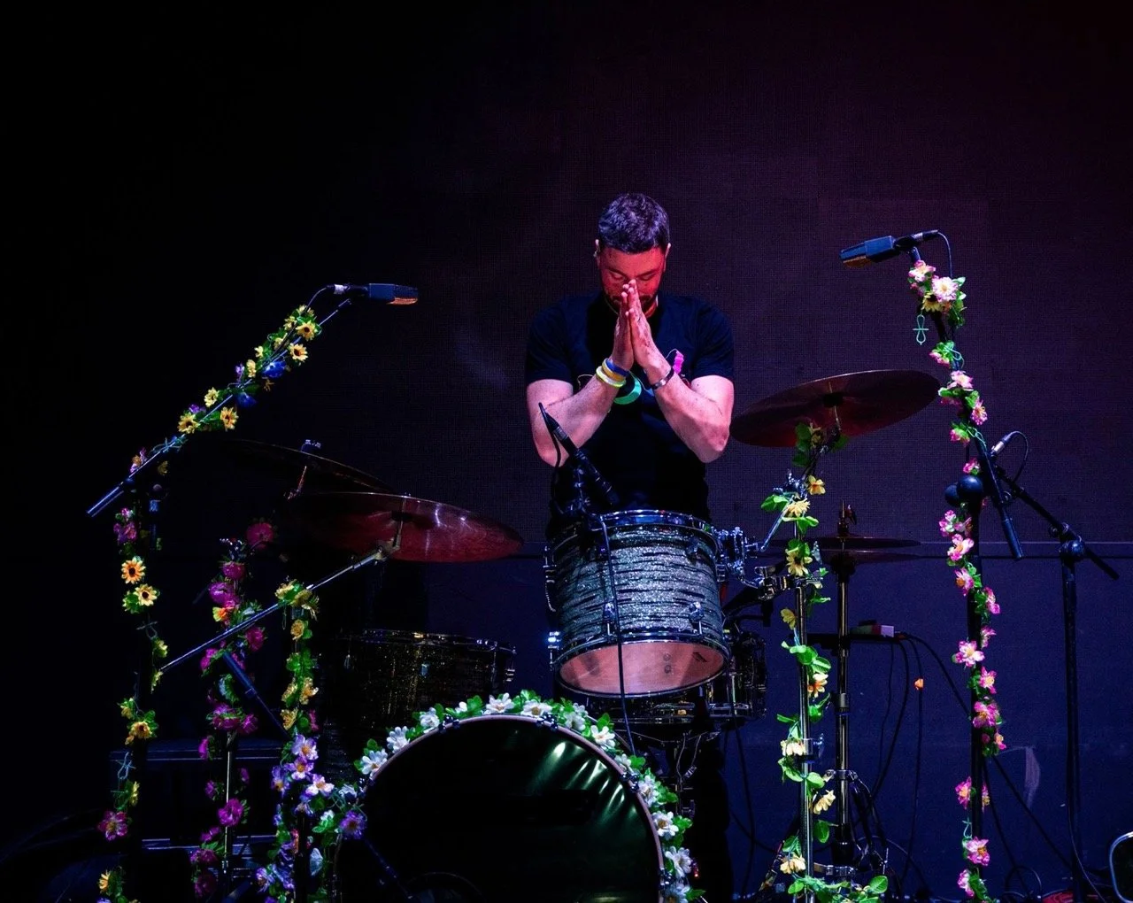 Musicista con mani giunte in preghiera, circondato da strumenti musicali decorati con fiori, sul palco durante un concerto.
