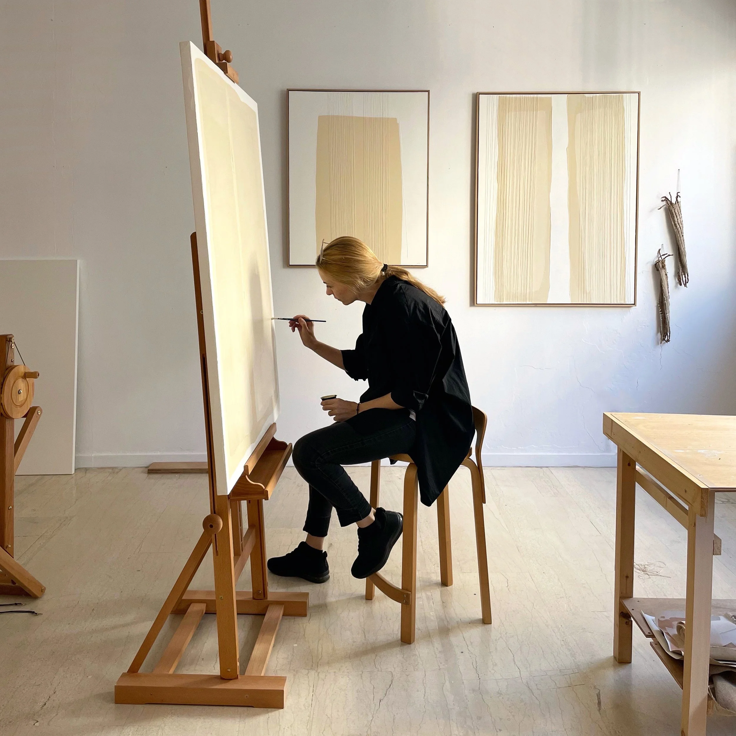 Visual artist Aggeliki Dimitriadou in her bright studio, seated at a wooden easel and painting on a large canvas, with two minimalist abstract artworks displayed on the white wall in the background.