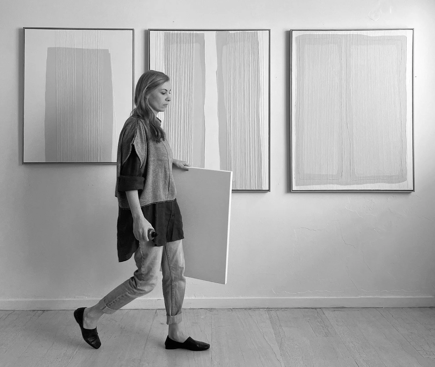 Black and white photograph of visual artist Aggeliki Dimitriadou in her studio. She is captured in motion, carrying a blank canvas in front of a white wall displaying three large-scale minimalist abstract paintings with vertical textures.