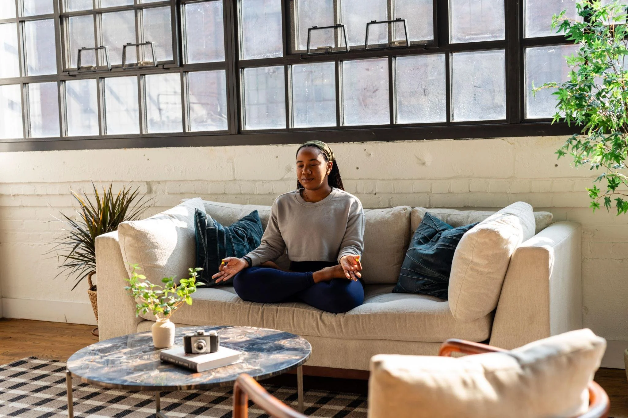A woman practicing meditation or mindfulness seated cross-legged on a sofa in a bright living room with large windows, plants, and a marble coffee table with books, a camera, and a small vase.