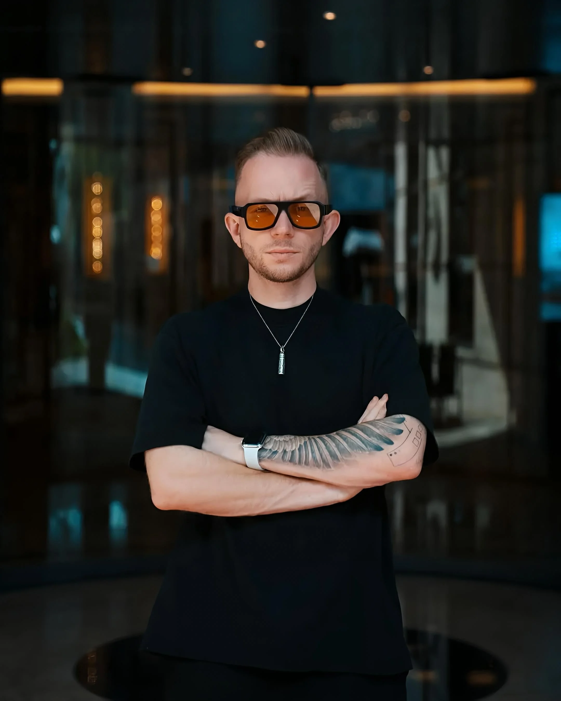 A man with short hair, beard, and tattooed arm wearing sunglasses, a black shirt, a necklace, and a watch, standing indoors with arms crossed, looking at the camera.