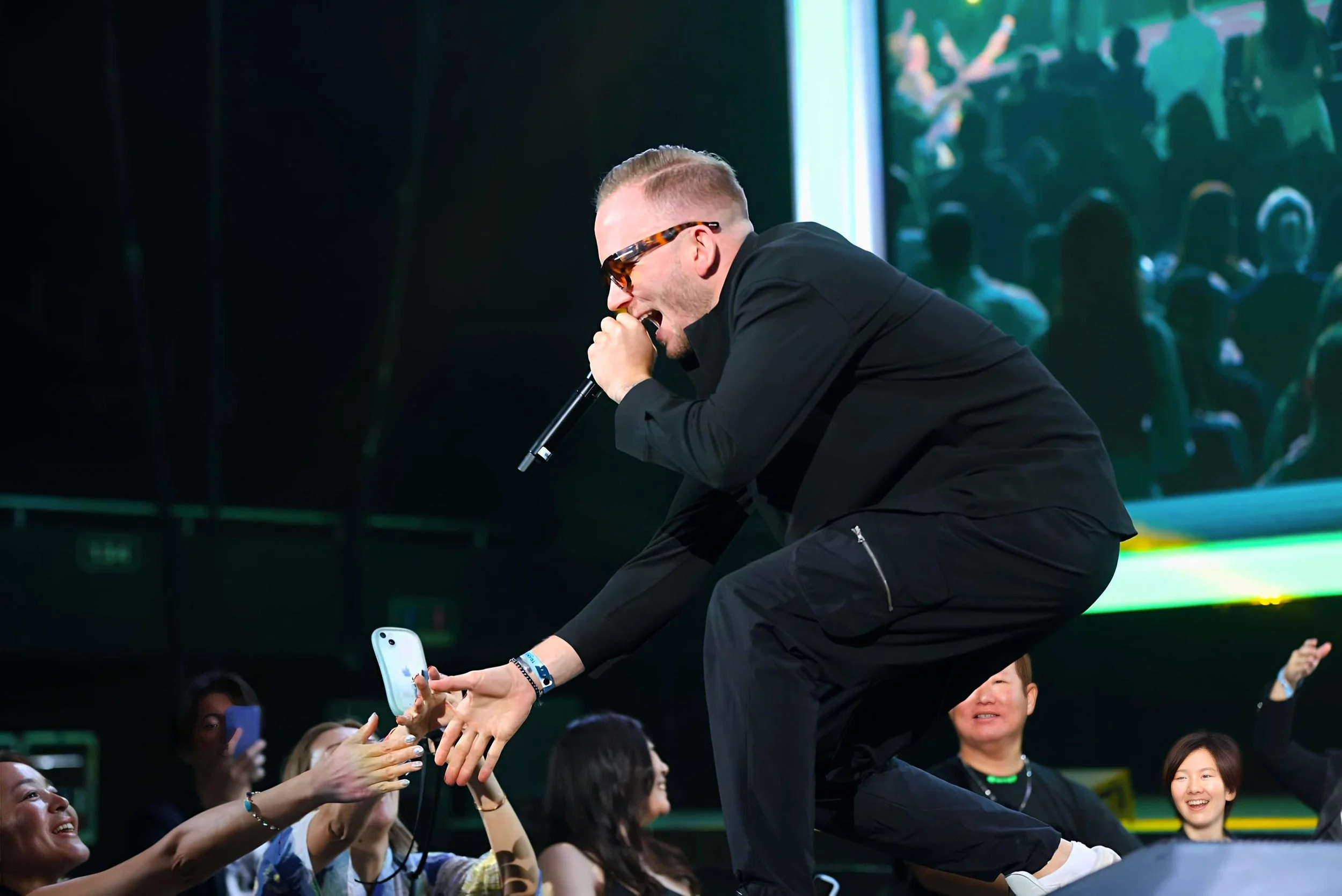 A male performer wearing sunglasses and black clothing is singing into a microphone while leaning over a crowd in an indoor venue. The crowd is reaching out to him, and some people are taking photos with their phones. The background shows a large screen with a colorful image and a semi-dark setting.