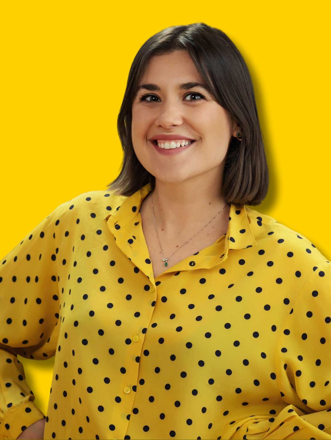 Smiling woman with shoulder-length dark hair wearing a yellow polka dot shirt against a yellow background.