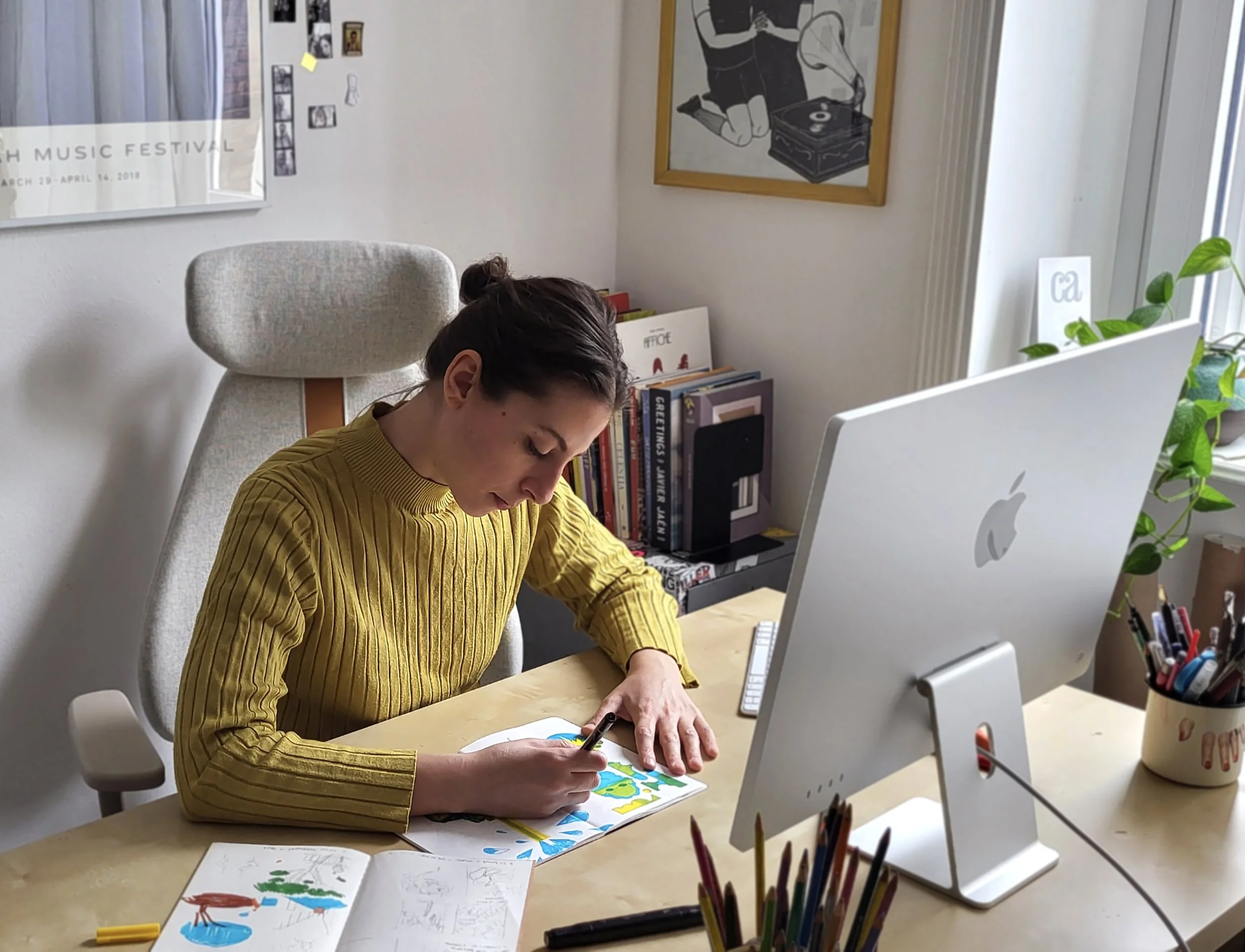 A woman with dark hair tied back, wearing a yellow striped shirt, is working at a desk with an iMac computer and colorful artwork in her hands. The background shows a white wall decorated with posters, photos, and a framed illustration of a person with a turntable.