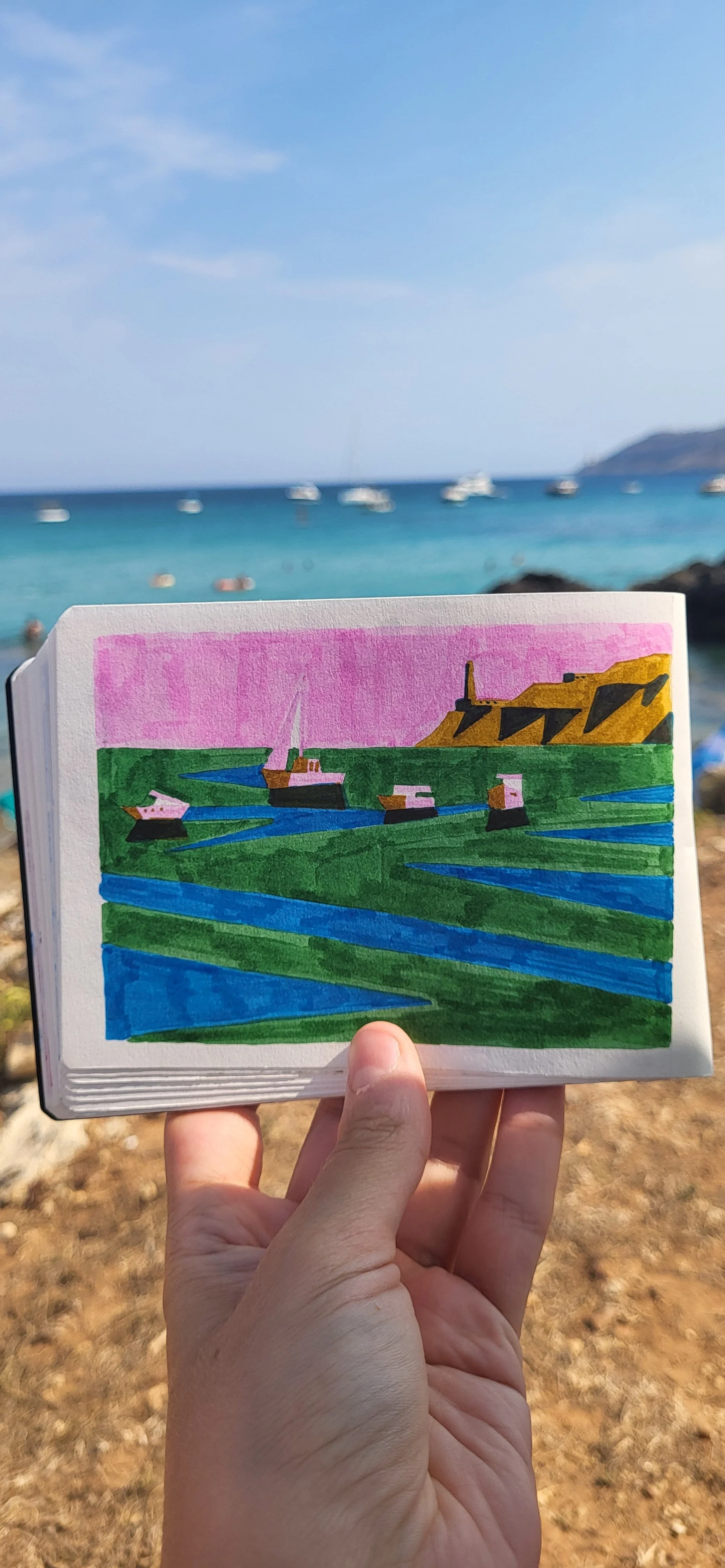 Photo of a hand holding a sketchbook with a colorful drawing of boats and cliffs, set against a beach with boats floating on blue water and a clear sky.