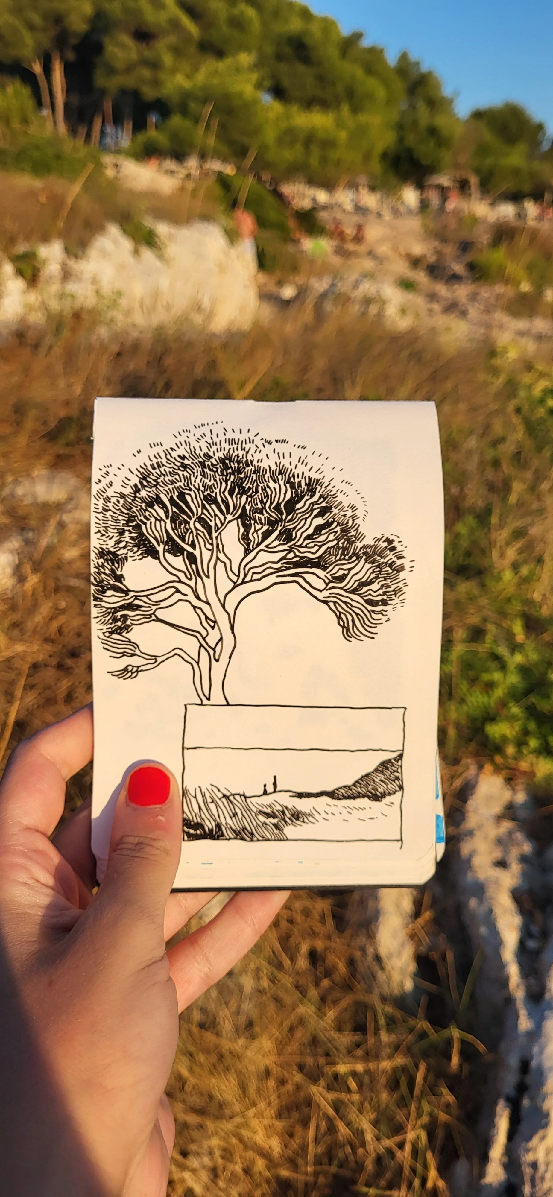 A person holding a black and white drawing of a tree with a person and a dog in a landscape, in front of a natural outdoor scene with grass, rocks, and trees.