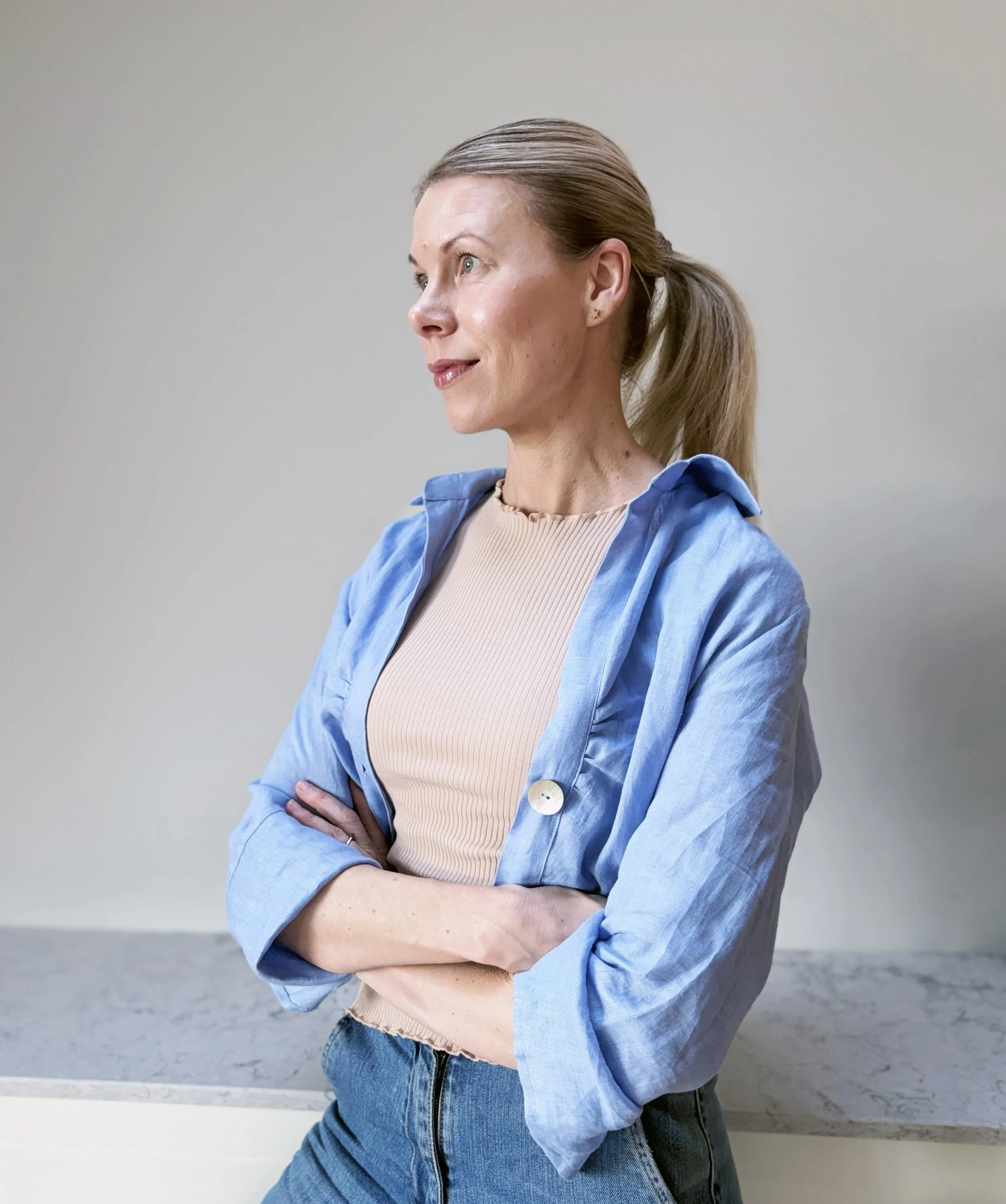 Kvinne med lyst hår i hestehale, iført beige topp, blå jakke og jeans står med armene i kors mot en enkel, lys vegg.
