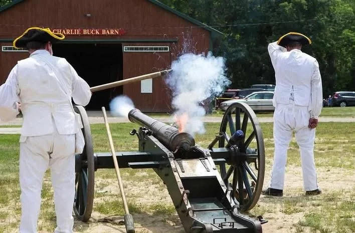Two men in white colonial uniforms firing a small black cannon outdoors, with one man holding a long stick and the other saluting, in front of a barn labeled "Charlie Buck Barn."