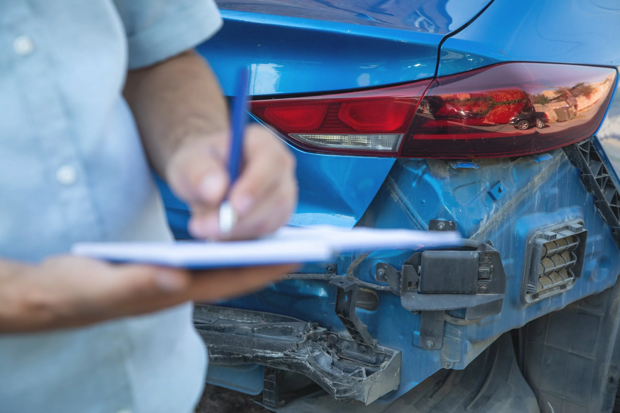 Ein blauer Auto vorne rechts mit Beschädigung, sichtbar durch die zerbrochene Stoßstange und beschädigte Front. Im Vordergrund eine Person, die einen Kuli hält, vermutlich bei der Dokumentation des Schadens.