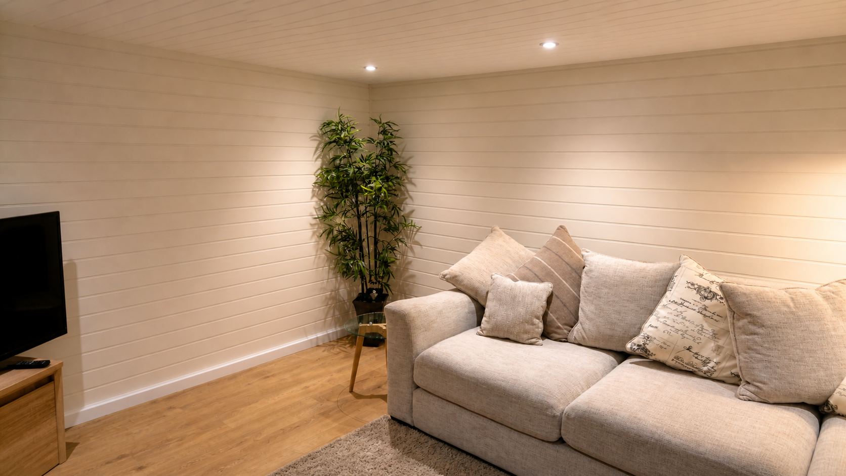 A cozy living room corner with a beige sofa decorated with multiple pillows, a tall greenhouse plant in the corner, a wooden TV stand with a television and remote, a small glass side table, and a wooden floor with a beige rug. The room has warm lighting and white paneled walls.