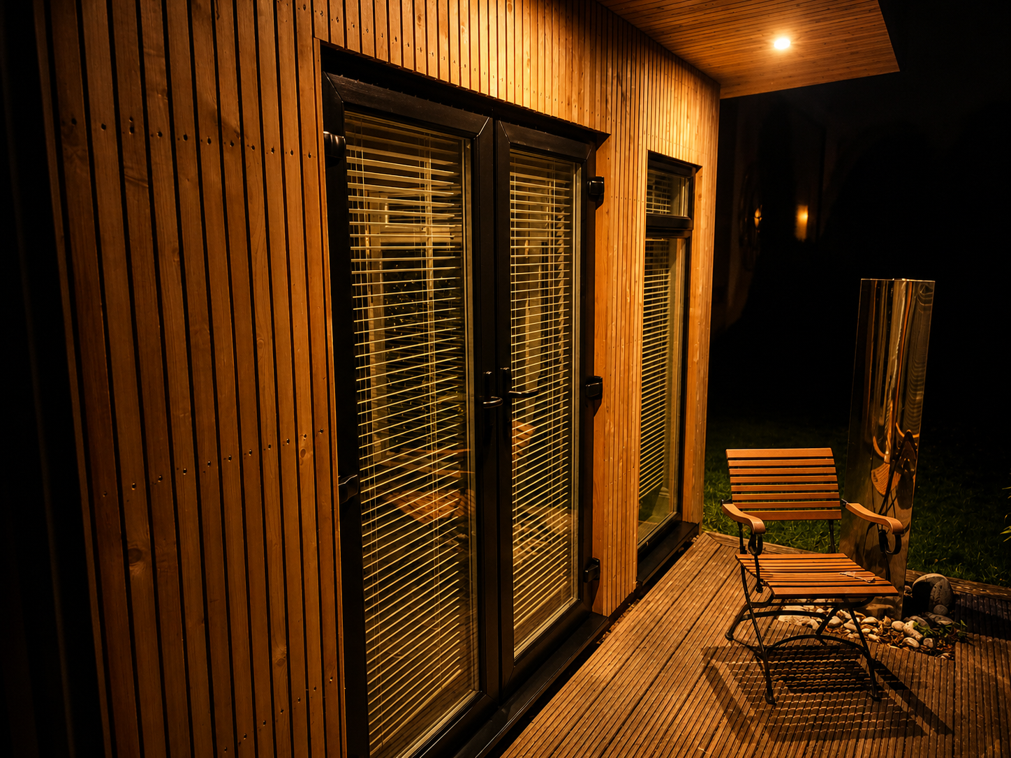 Nighttime view of a wooden deck outside a house with sliding glass doors and blinds, illuminated by a ceiling light, with a wooden chair and a metallic sculpture on the deck.