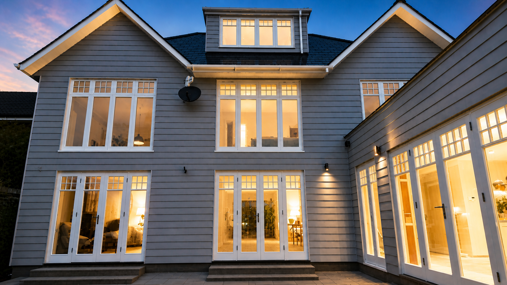 Modern multi-story house with large windows, during dusk, with warm interior lighting visible through the glass doors and windows.