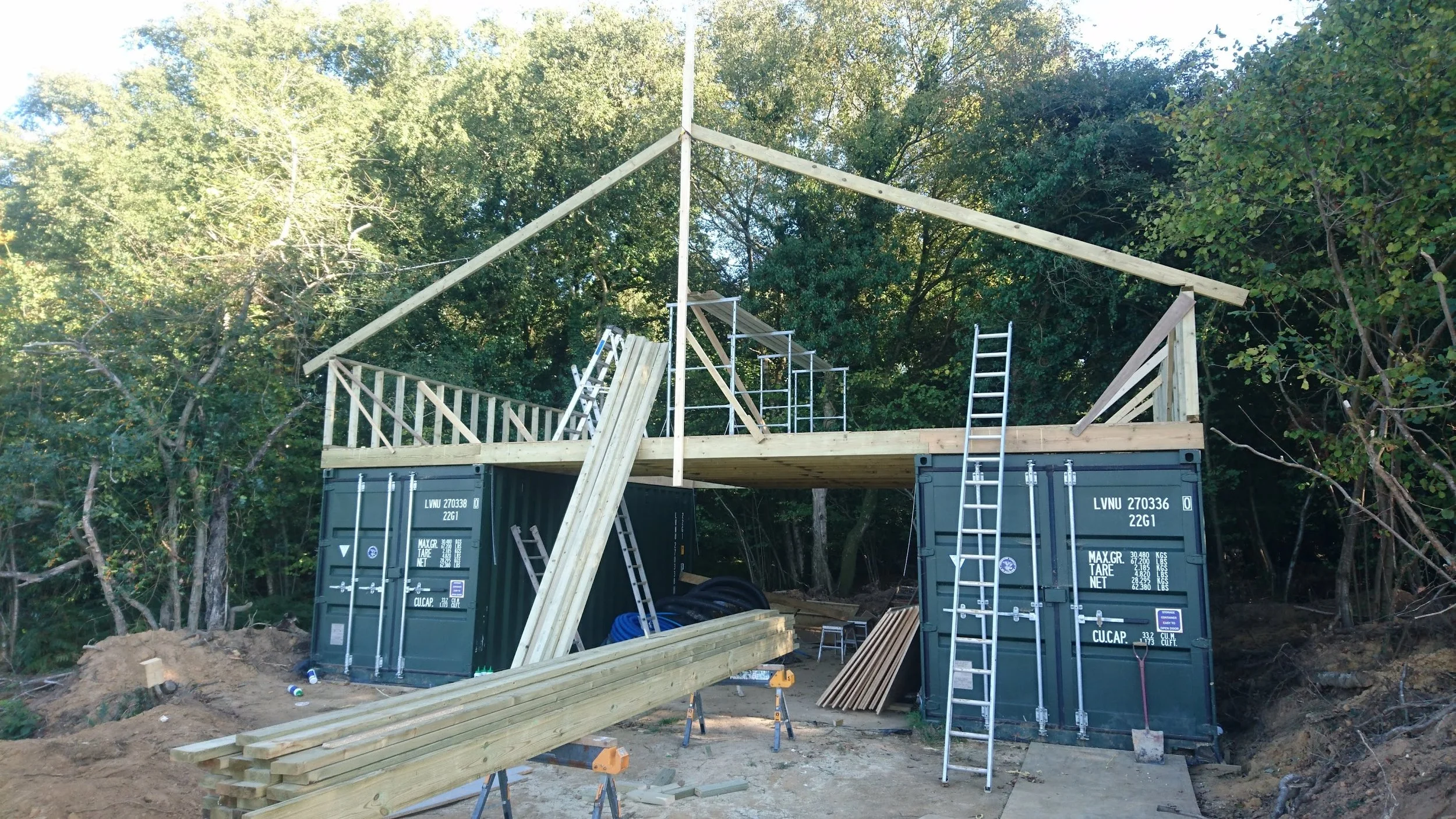 Construction site with a wooden house frame being built on top of a shipping container, surrounded by trees.