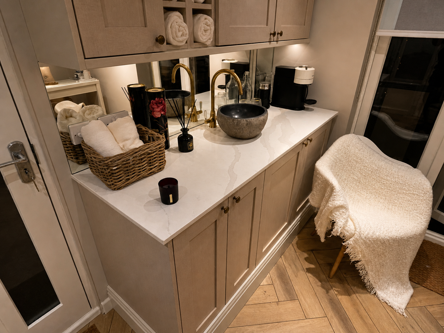 A bathroom vanity with a white marble countertop, a stone vessel sink, and gold fixtures. Items include rolled towels, black candles, a reed diffuser, a black coffee machine, and glassware. A wicker basket with towels sits on the counter, and a cozy white armchair with a fringed throw is nearby. There are wood cabinets above and below, and a sliding glass door to the right.