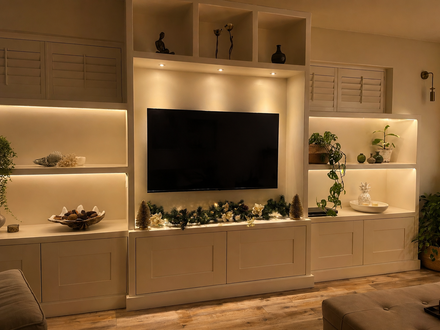 Living room entertainment center with a large television, holiday decorations, potted plants, and shelves with decorative items under warm lighting.