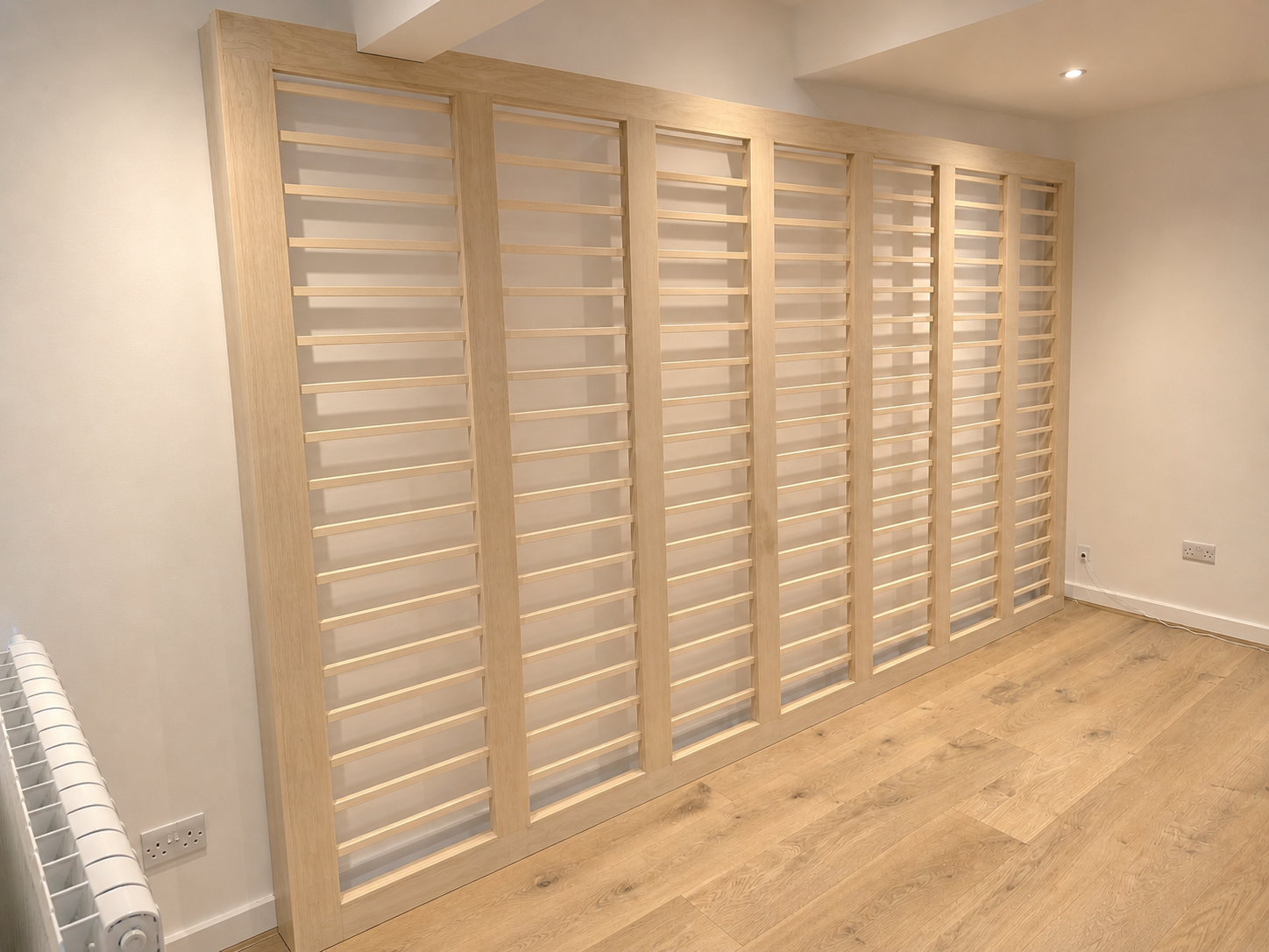 Unfinished wooden wardrobe with multiple slatted doors in a room with light-colored walls and hardwood flooring.