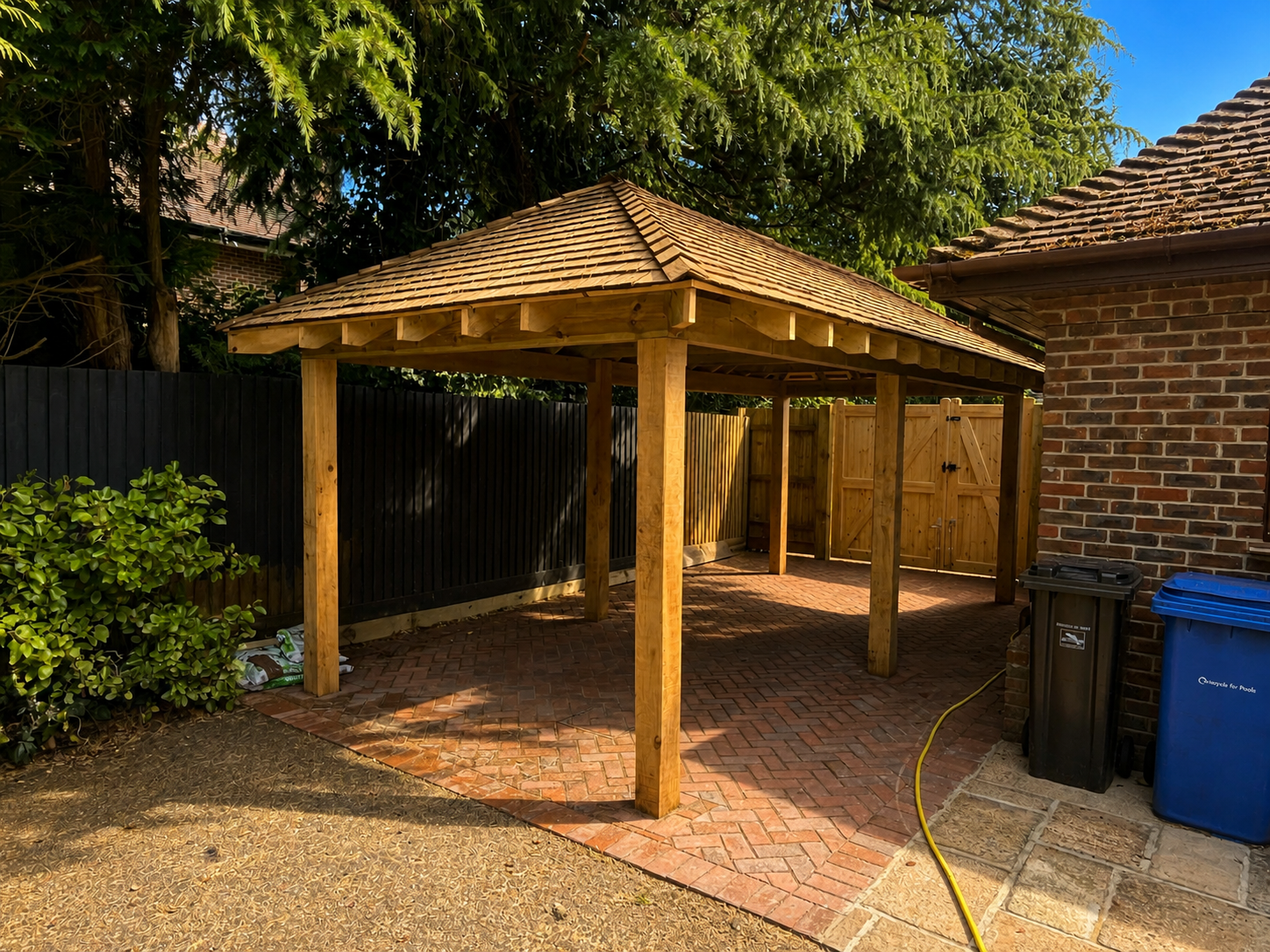 Wooden gazebo with a shingled roof in a backyard, brick-paved patio, black fence, brick house wall, two garbage bins, and a yellow garden hose.