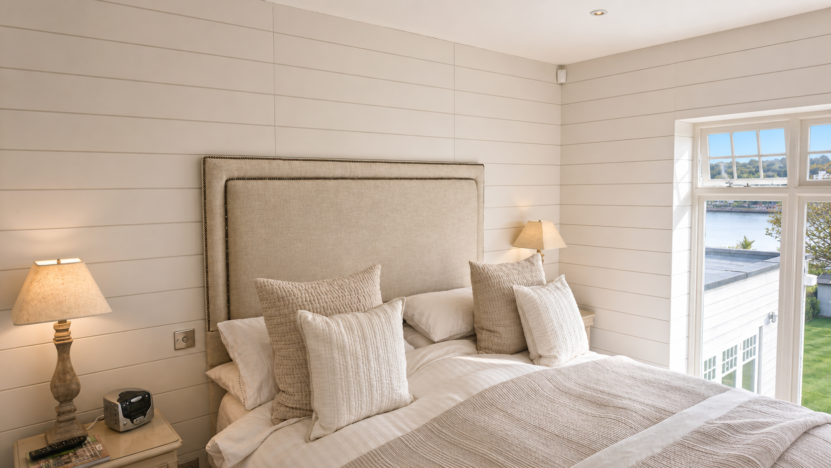 A cozy bedroom with a large beige upholstered headboard, multiple cream-colored pillows, and beige bedding. There are two bedside lamps with beige lampshades, a small alarm clock, and magazines on the nightstand. A window with multiple panes shows a view of a river, trees, and houses outside.